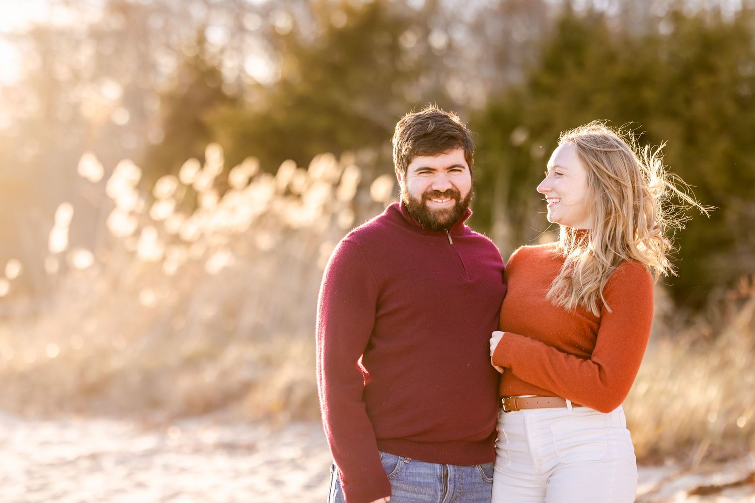 Couple laughing together in burgundy and orange sweaters during golden hour.