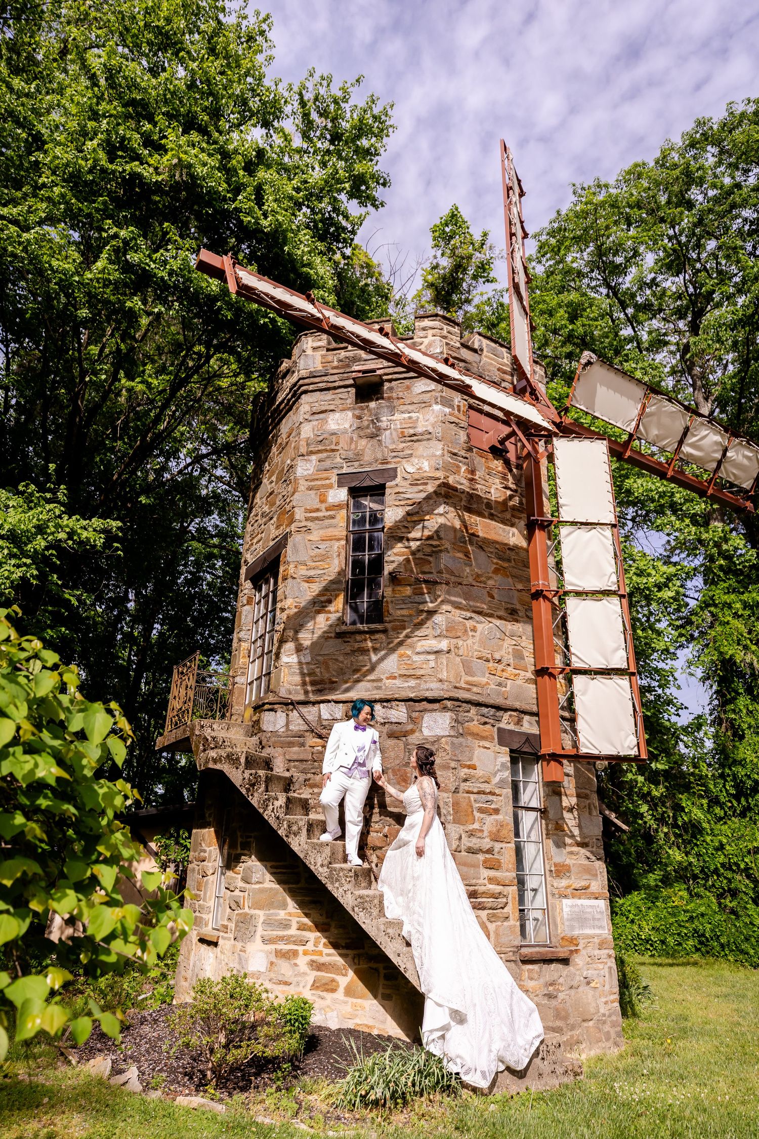 A wedding couple poses on the steps of a rustic stone windmill or tower structure in a garden setting.
