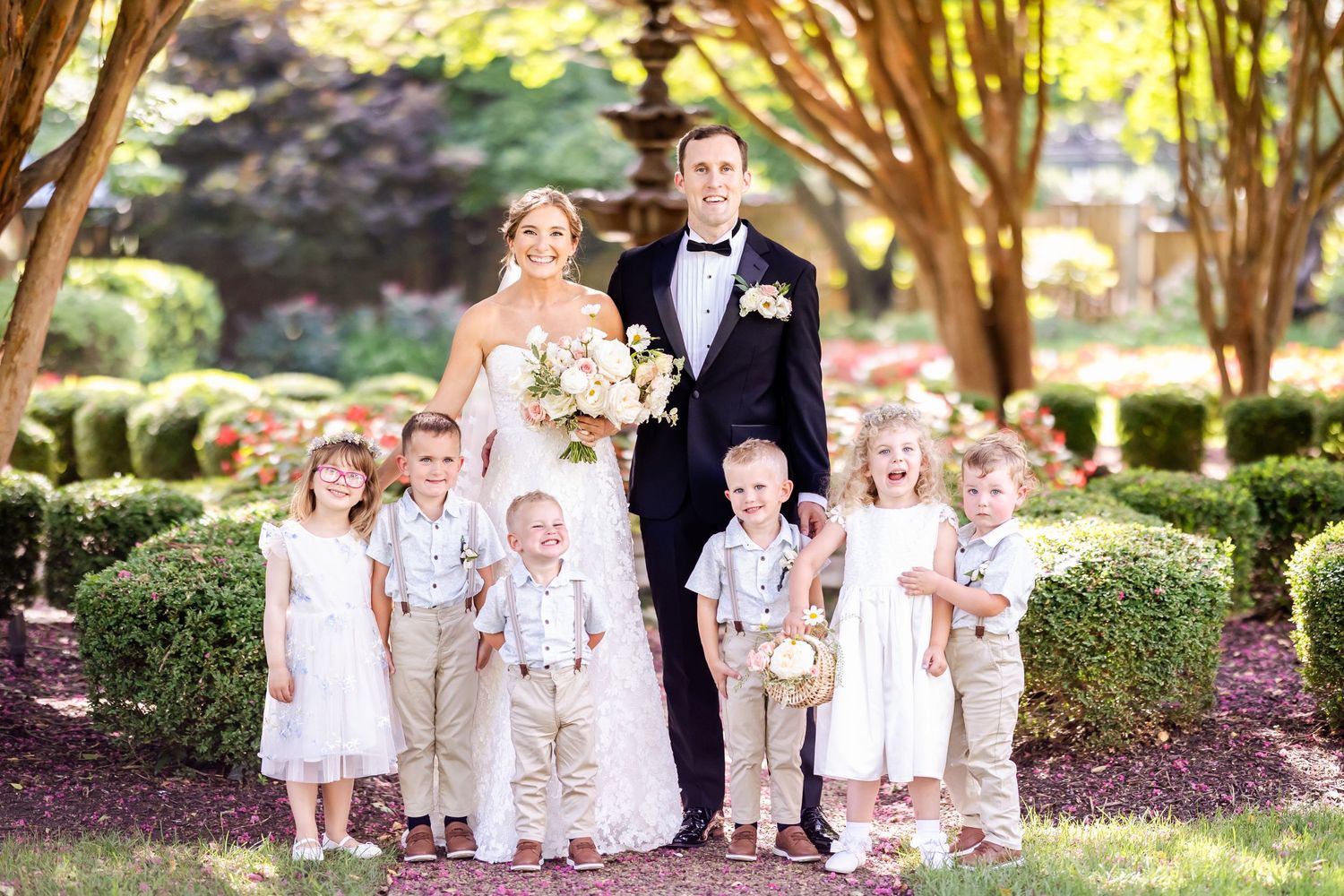 A wedding party poses outdoors with a bride and groom accompanied by six young children in formal attire.