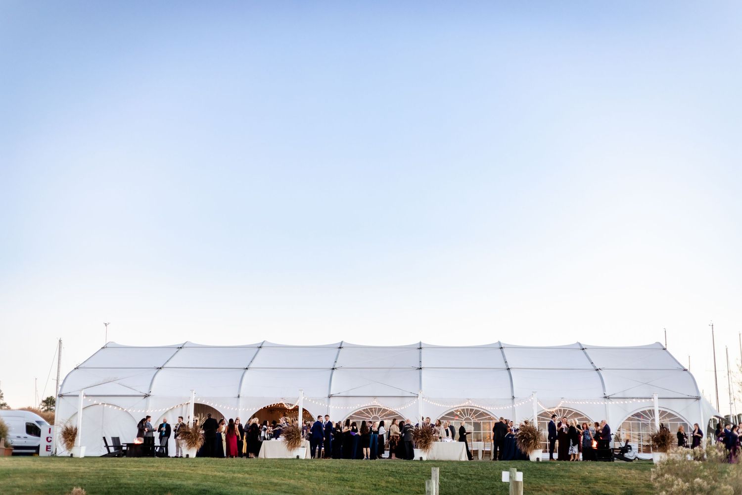 Large white tent pavilion set up on grass for an outdoor wedding reception with guests gathering at sunset.