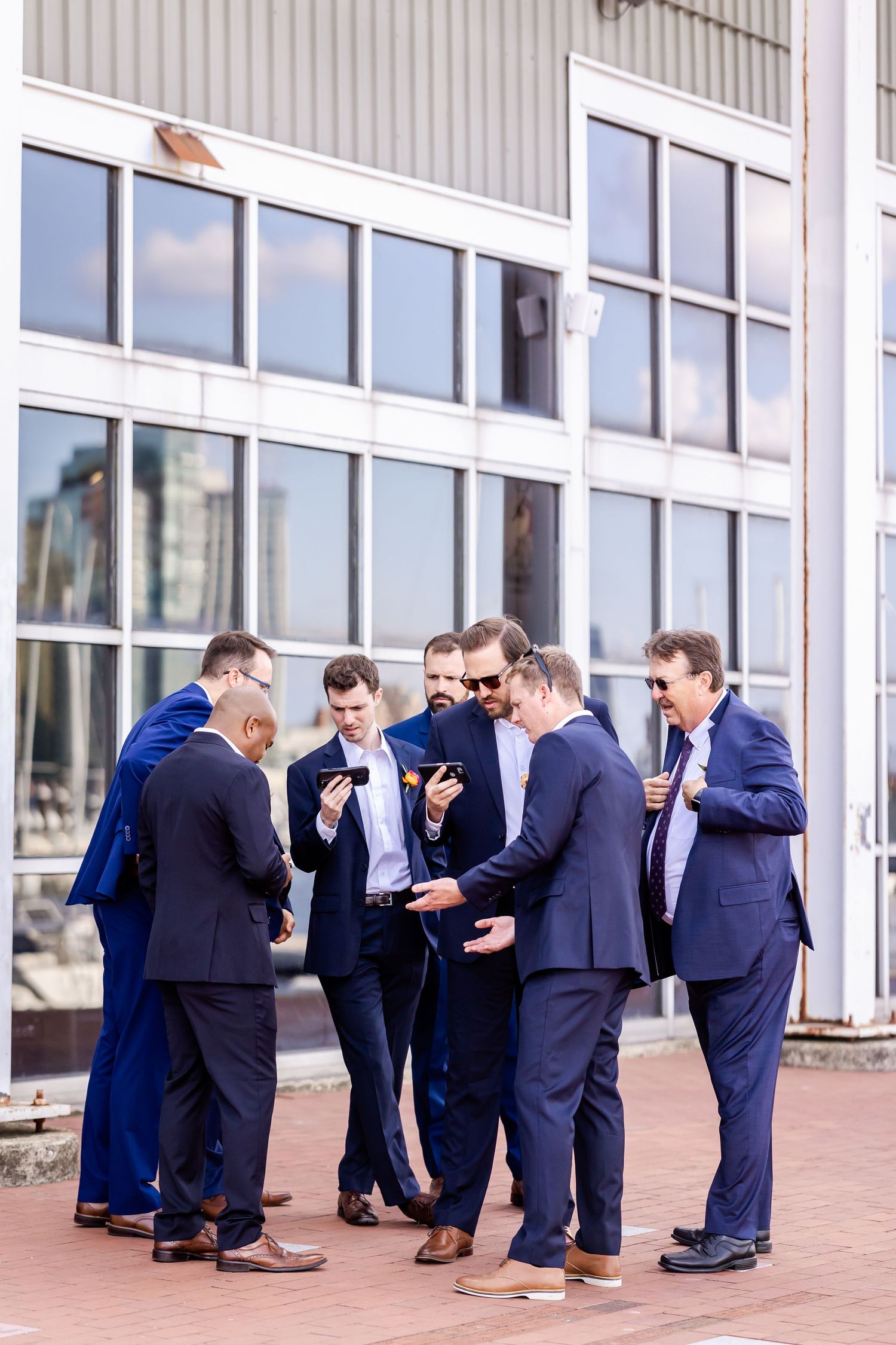 Groomsmen in navy suits gathered in circle on brick patio looking at something together.