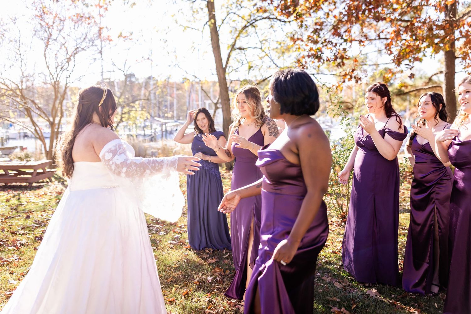 Bride in white gown shares special moment with bridesmaids in purple dresses during outdoor autumn ceremony.
