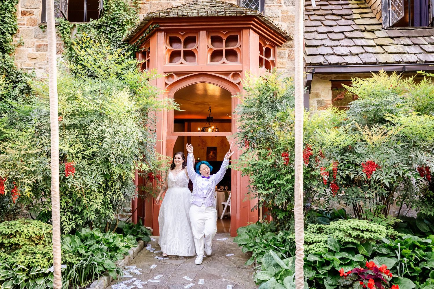 Two people in white attire walk through an ornate terracotta-colored archway surrounded by flowering gardens.