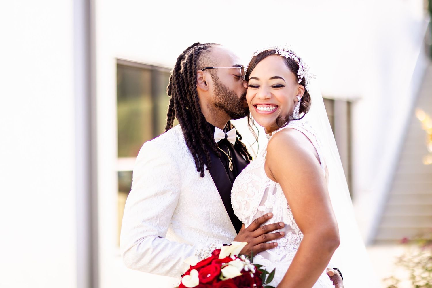 Happy couple laughing together in elegant wedding attire with red rose bouquet.