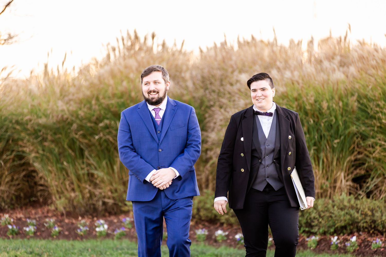 Two gentlemen in contrasting blue and black formal suits stand in front of tall ornamental grasses.
