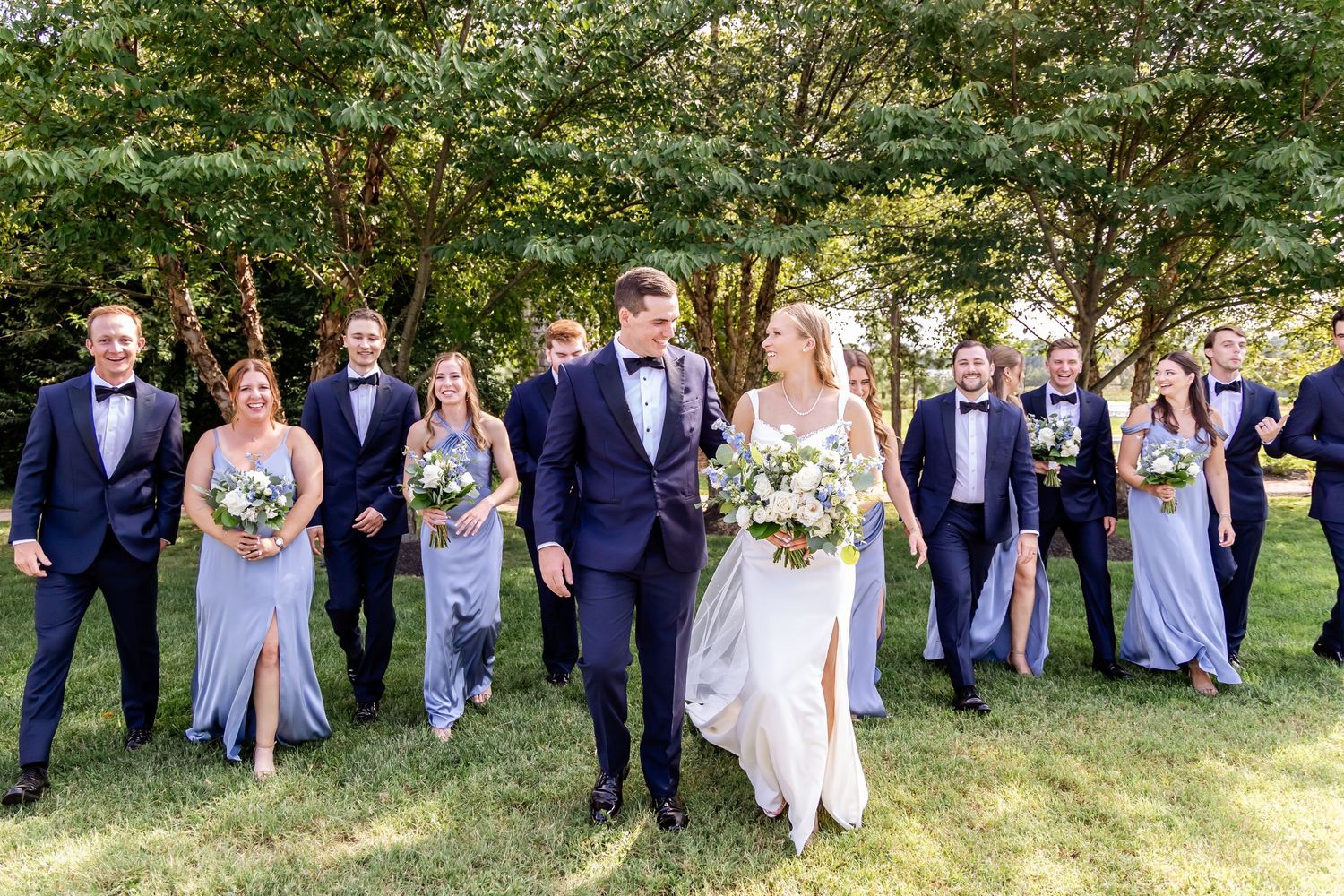 Wedding party in navy suits and light blue dresses pose together on grassy lawn for photo during beautiful outdoor ceremony.