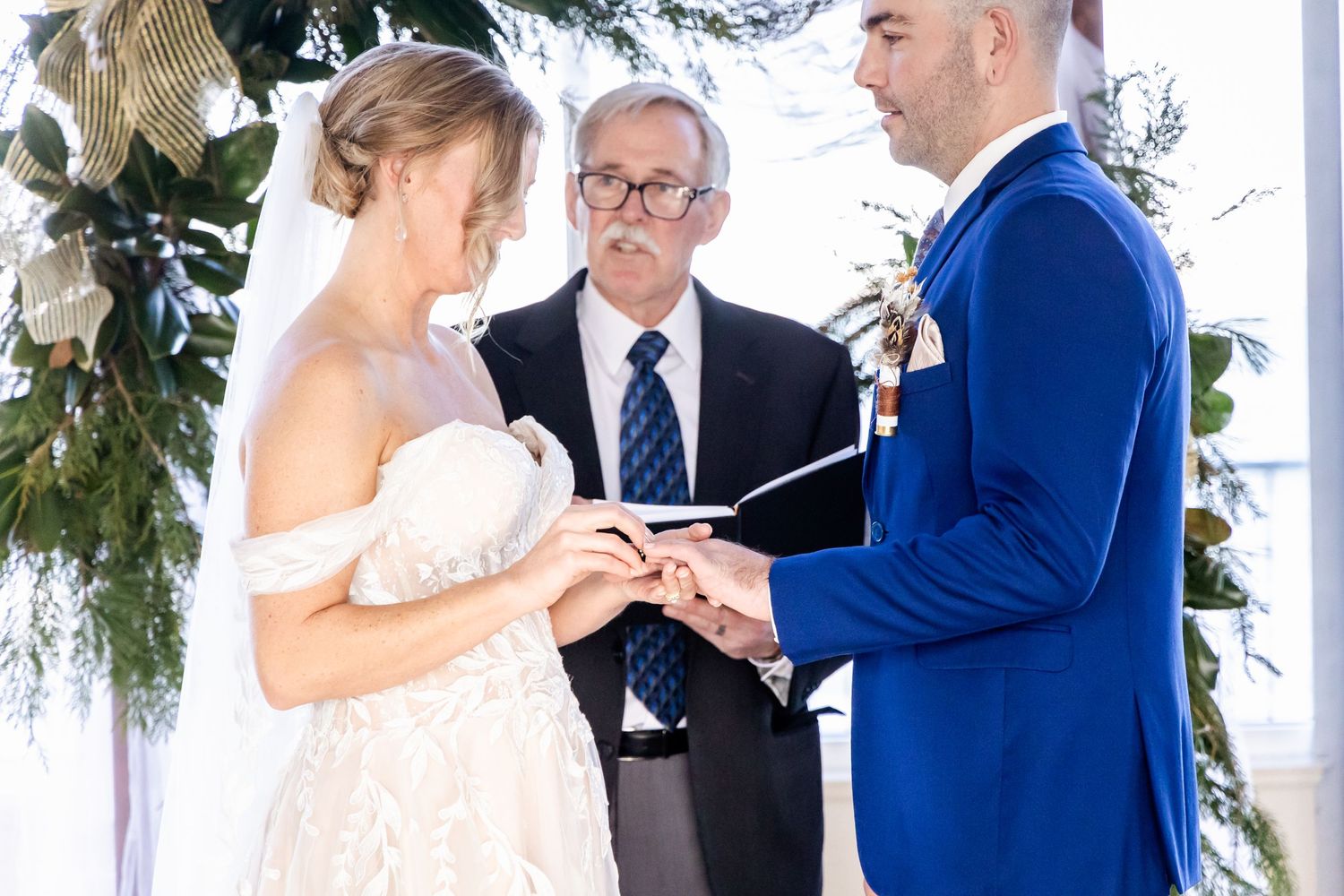 Wedding ceremony with couple exchanging rings under greenery arch decorations.