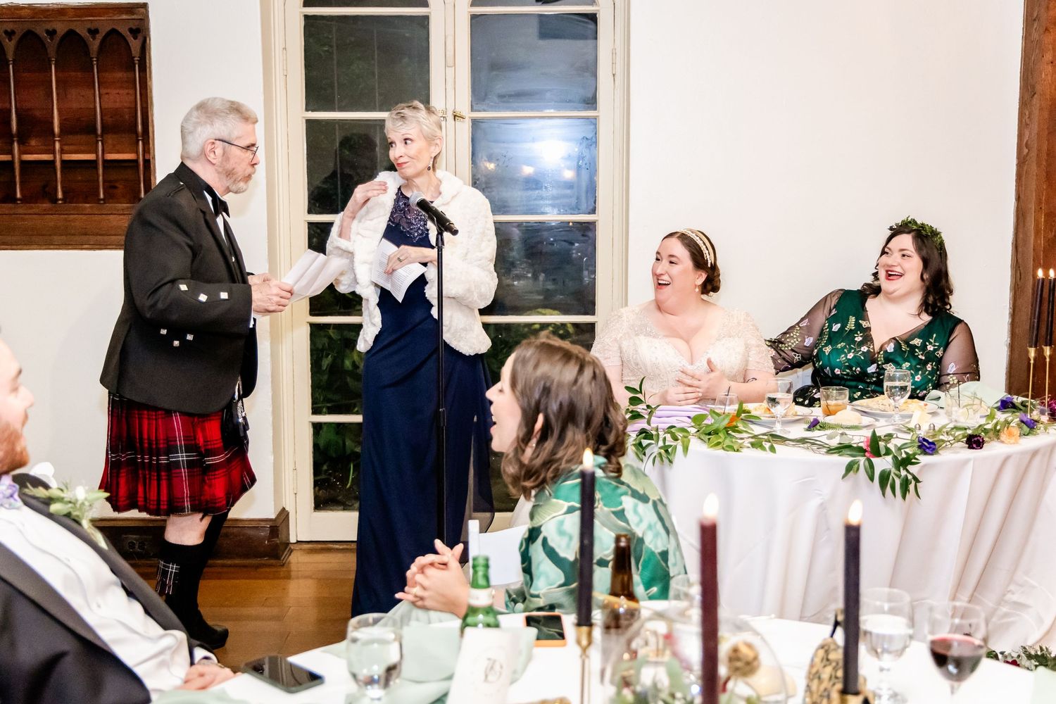 A lively wedding reception with guests at a banquet table while a man in a kilt makes a toast.