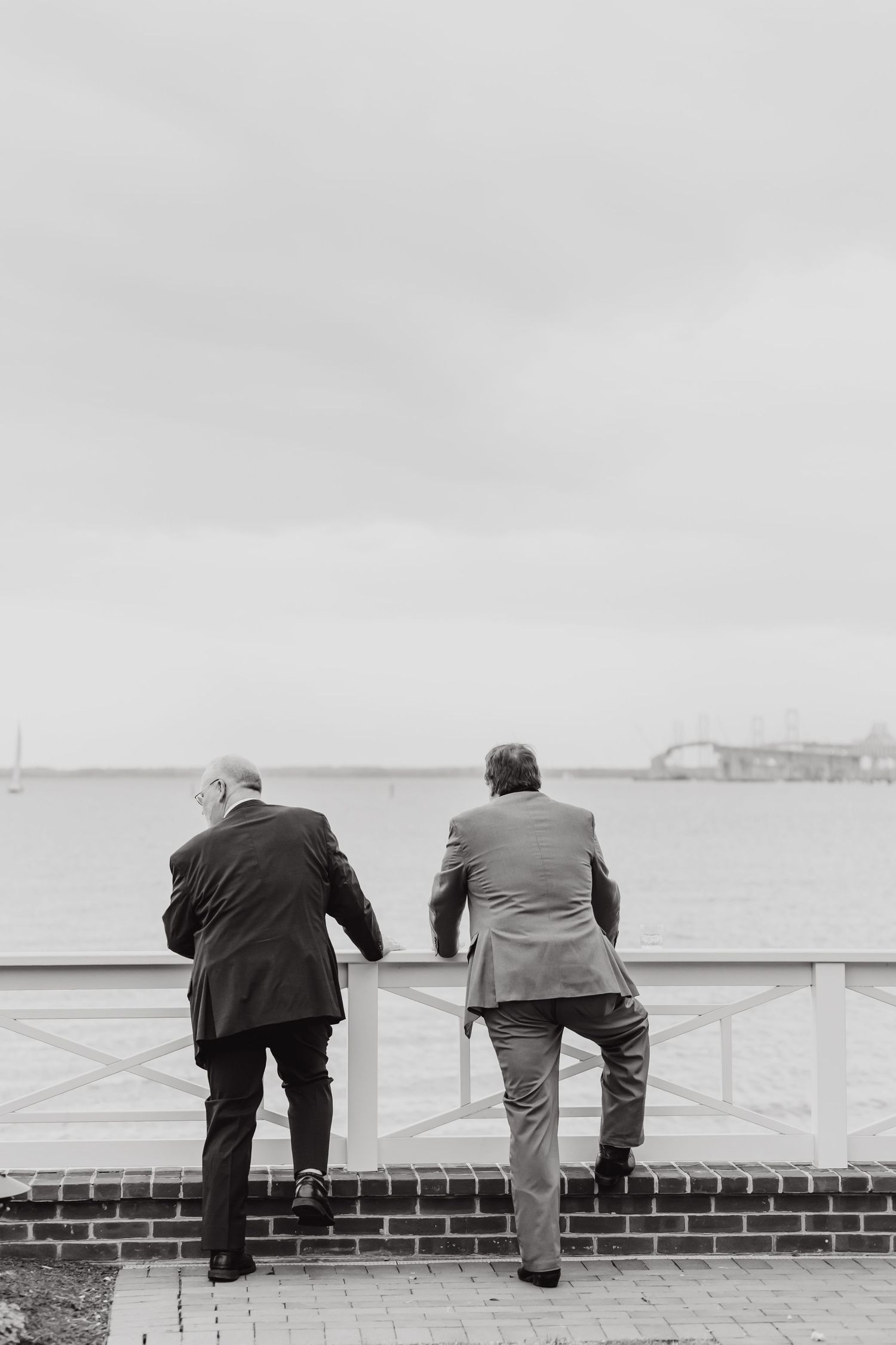 Two figures in suits lean against a railing overlooking a waterfront view in black and white.