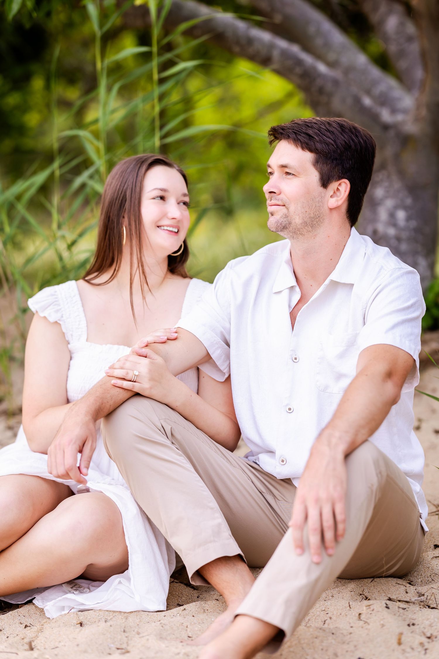 A romantic couple in white clothing sits together on a sandy beach with lush greenery in the background.