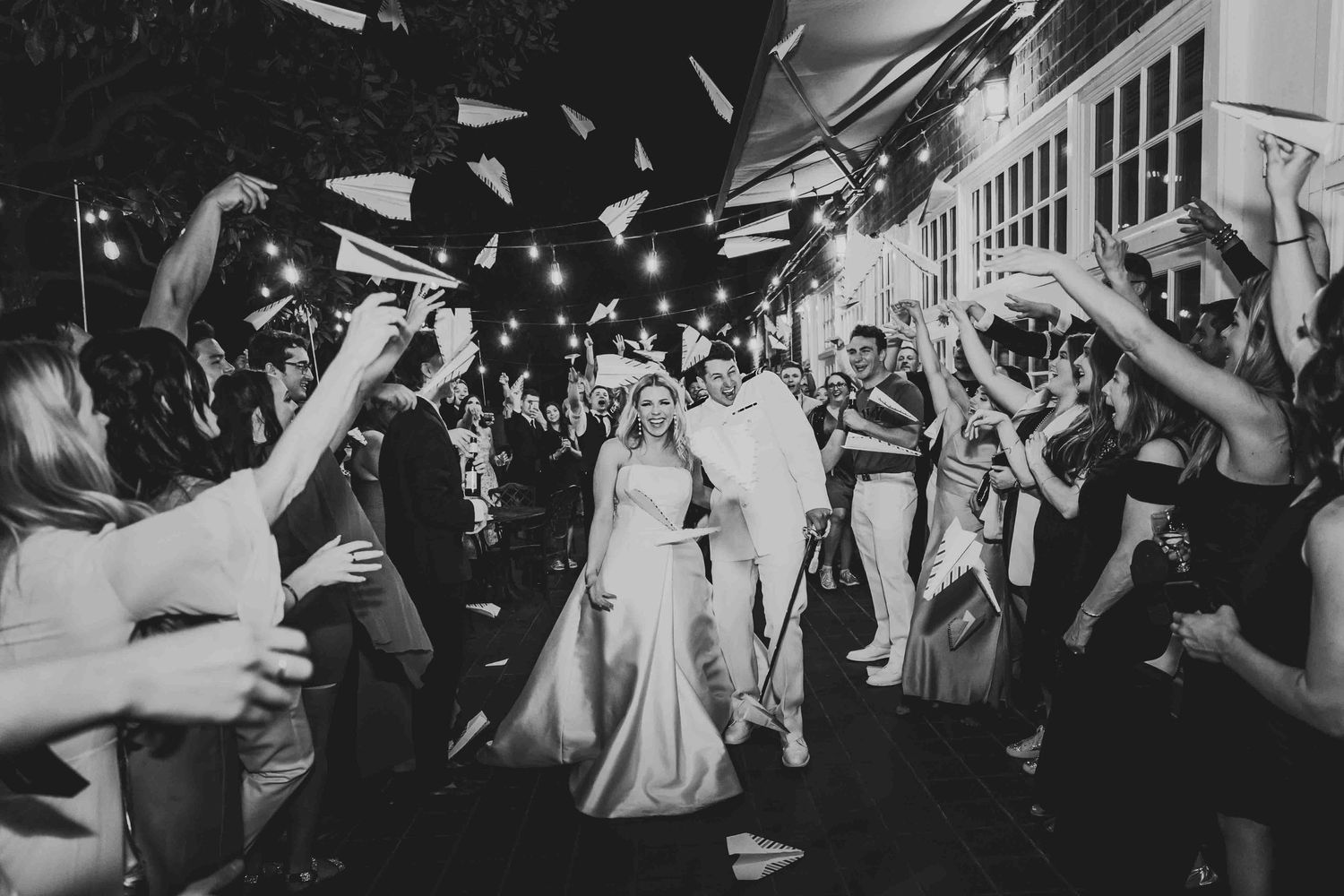 Wedding guests celebrate with bubbles at night as couple leaves their reception in black and white photography.