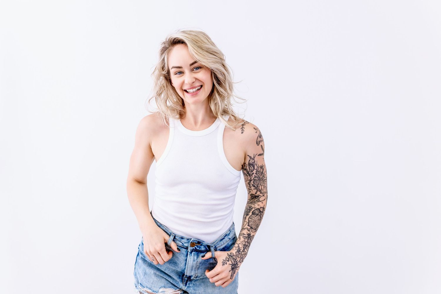 Person wearing white tank top and denim shorts poses casually against white background showing off sleeve tattoos.