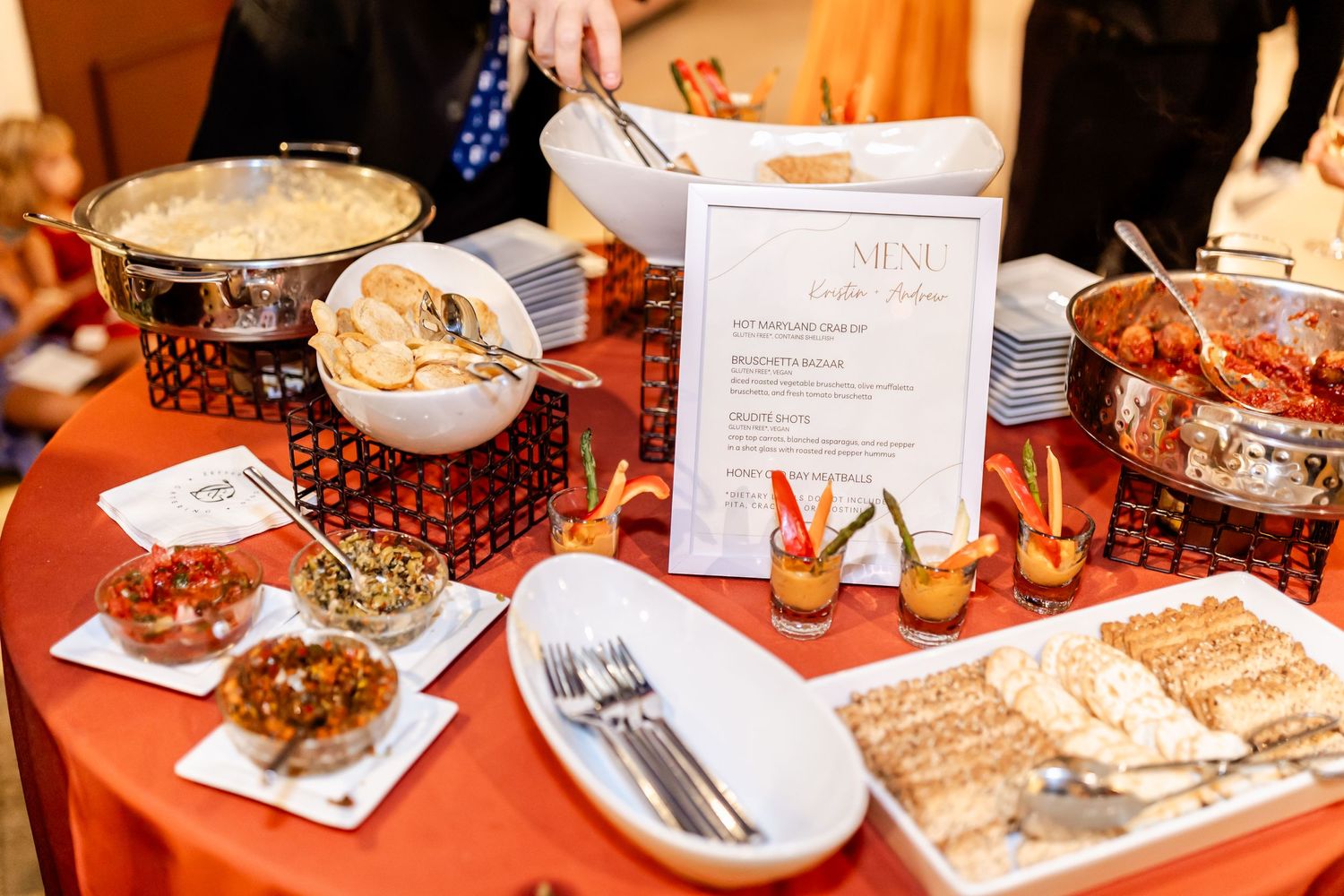 Catering table set with appetizers and menu cards on orange tablecloth.