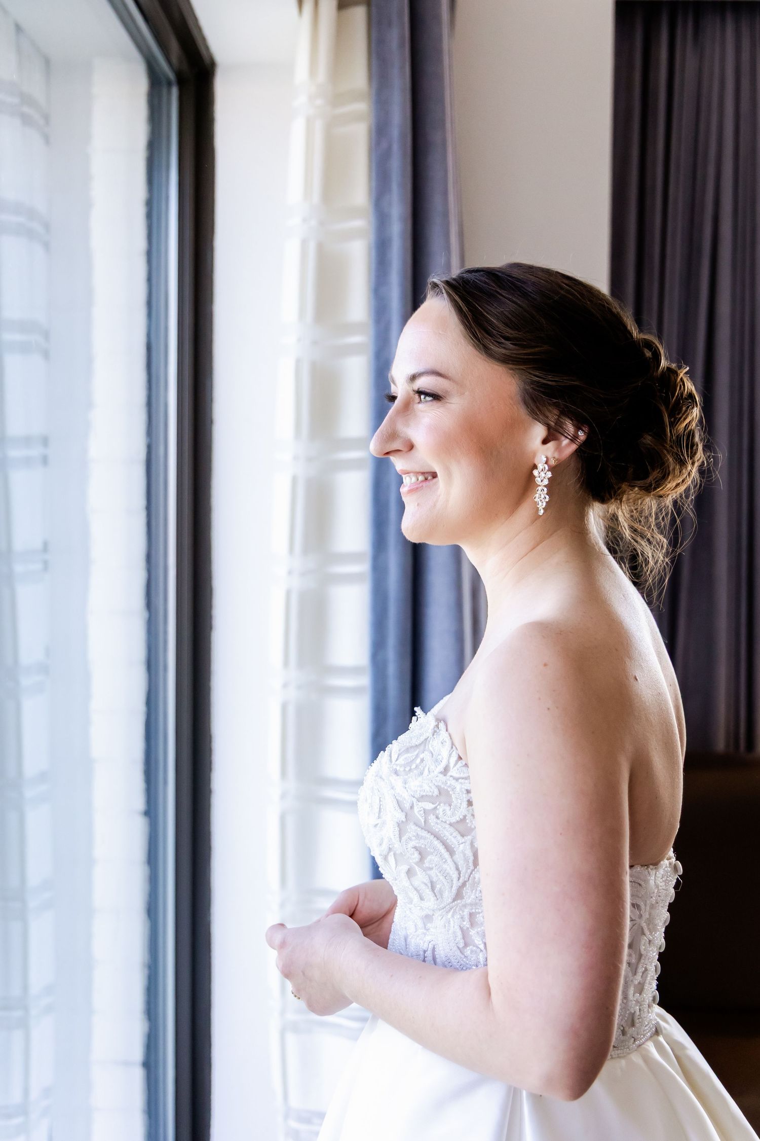 Beautiful bride in a lace strapless wedding gown smiles while looking out a window with elegant curtains.