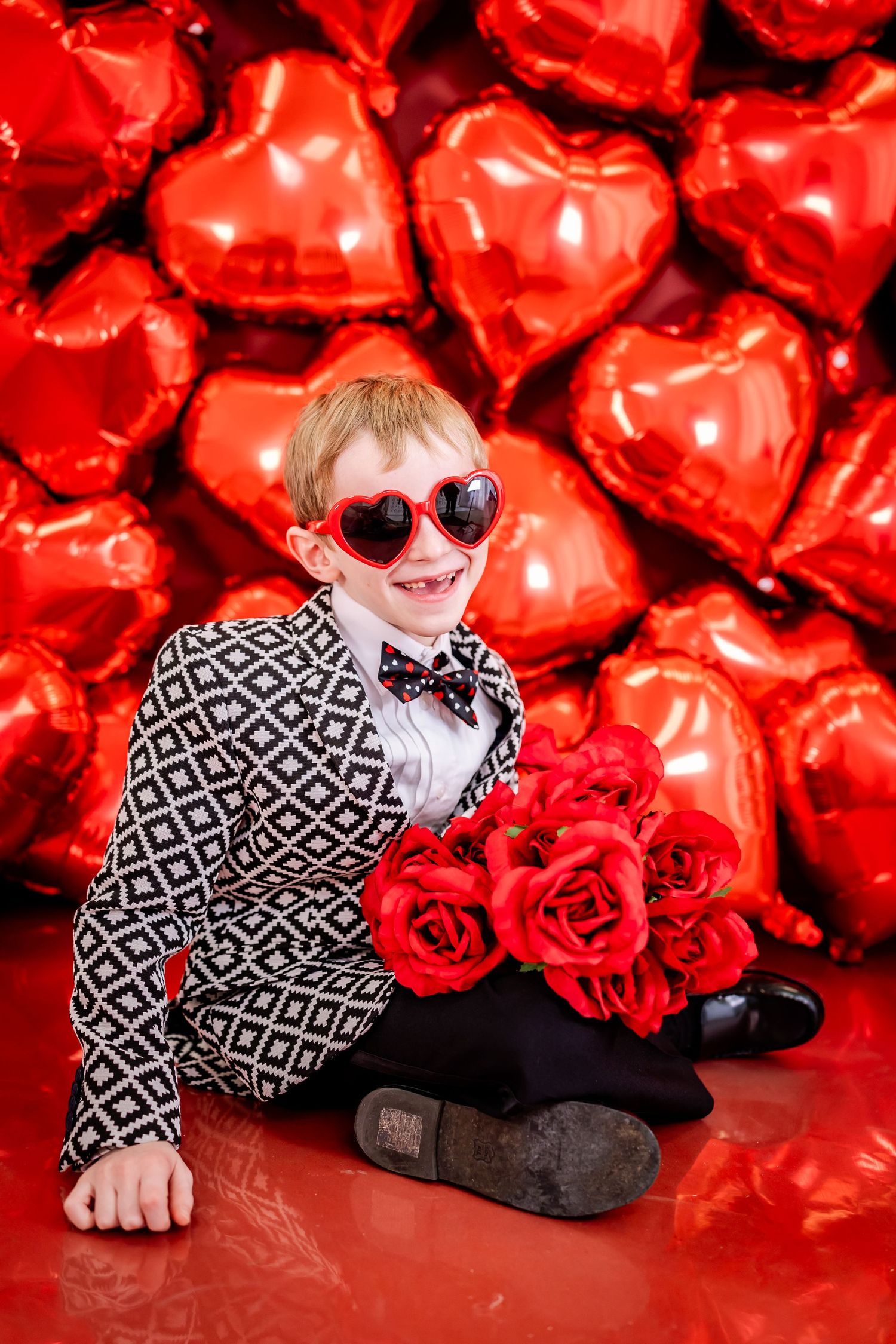 Stylish person in geometric pattern suit and heart sunglasses poses with red roses against backdrop of heart balloons.