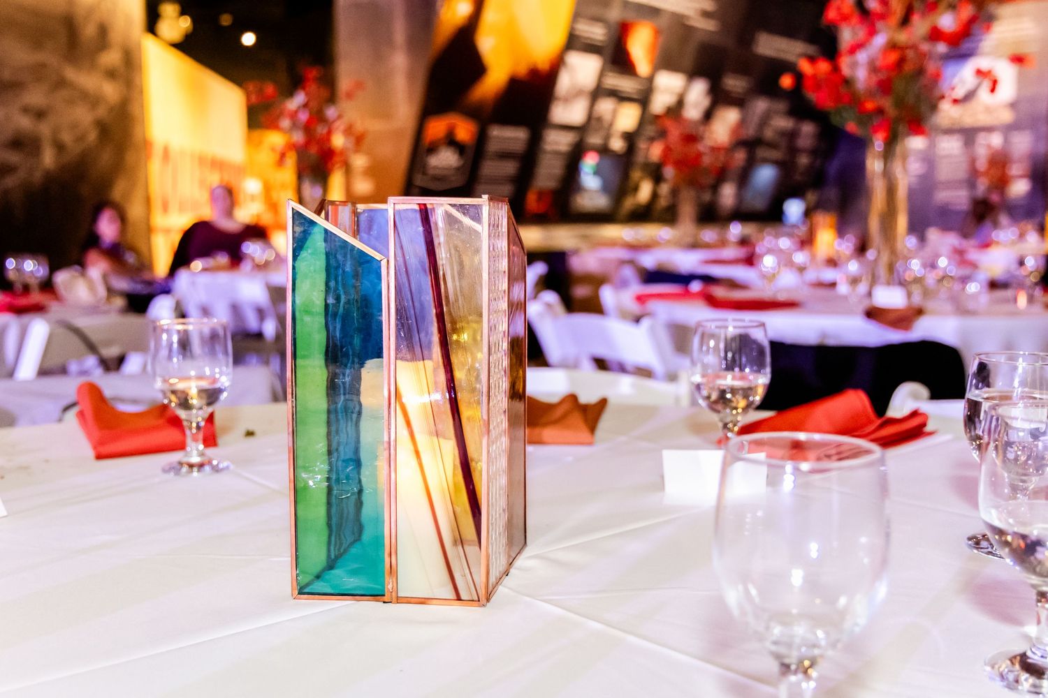 A colorful stained glass table centerpiece glows on a formal dining table with wine glasses and red napkins.