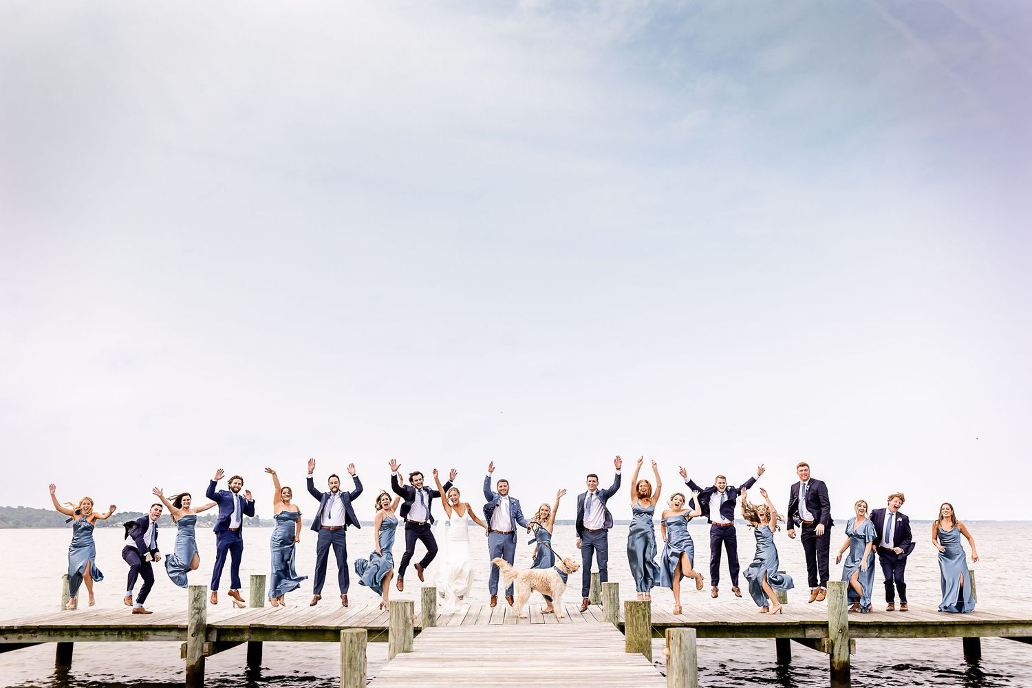 Wedding party celebrates with raised arms on a pier against a bright white sky and ocean backdrop.