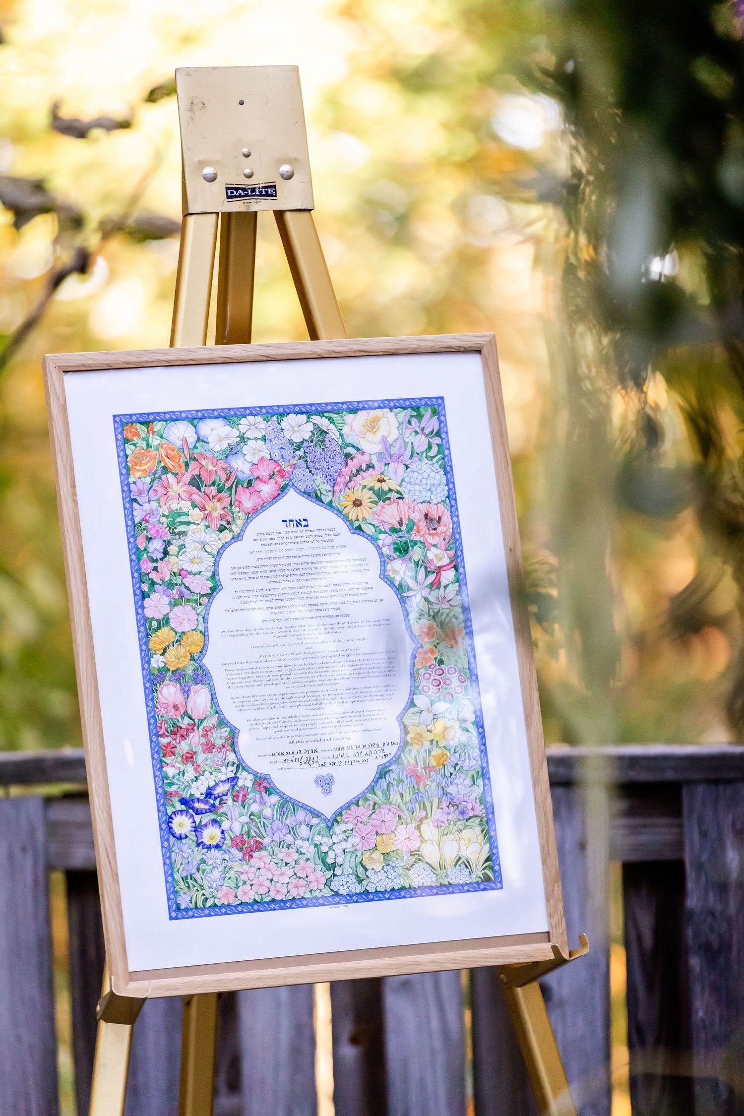 Framed floral wedding ketubah displayed on a wooden easel in soft outdoor lighting.