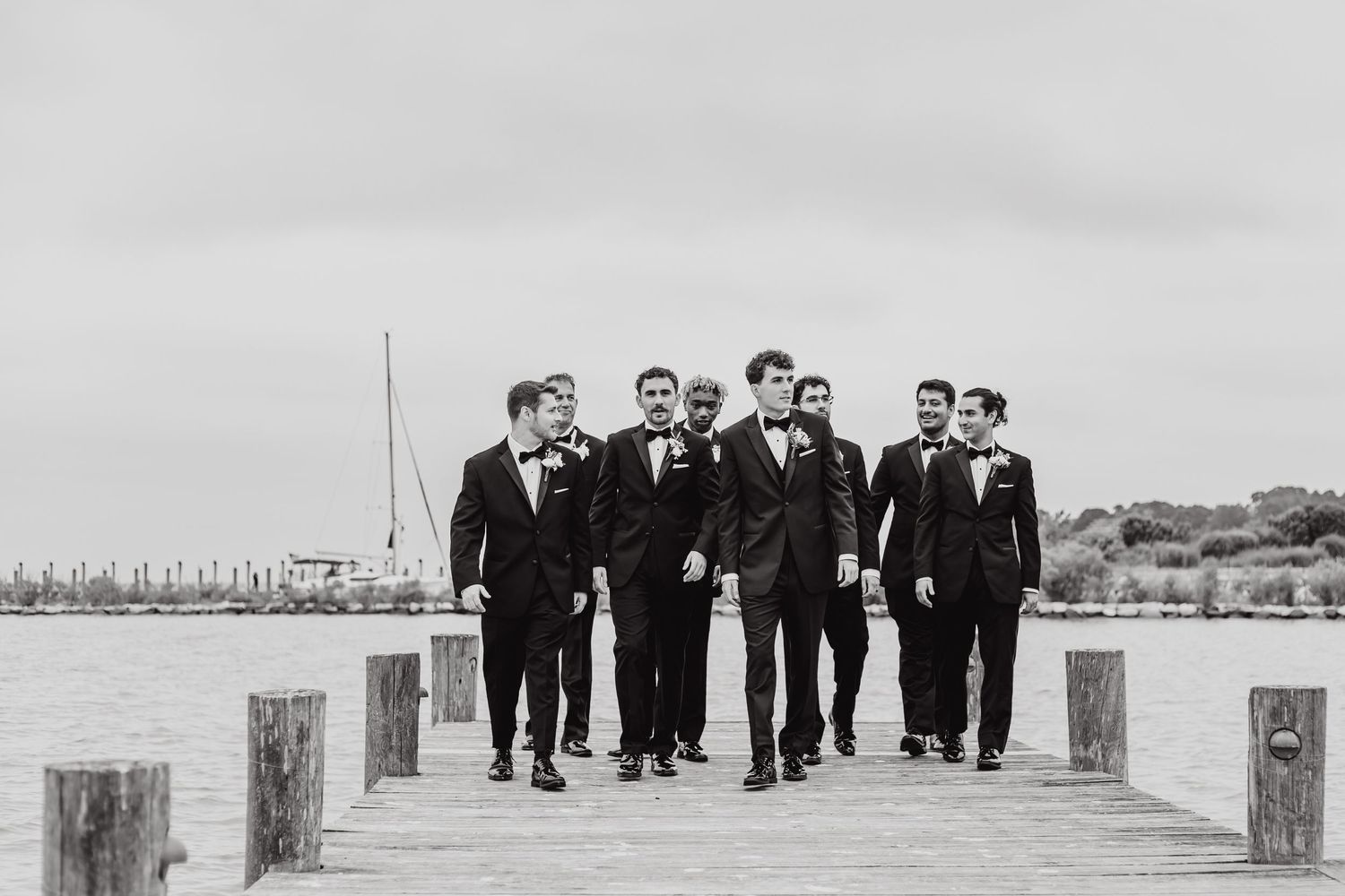 Groomsmen walking along a wooden pier by the water in black and white.