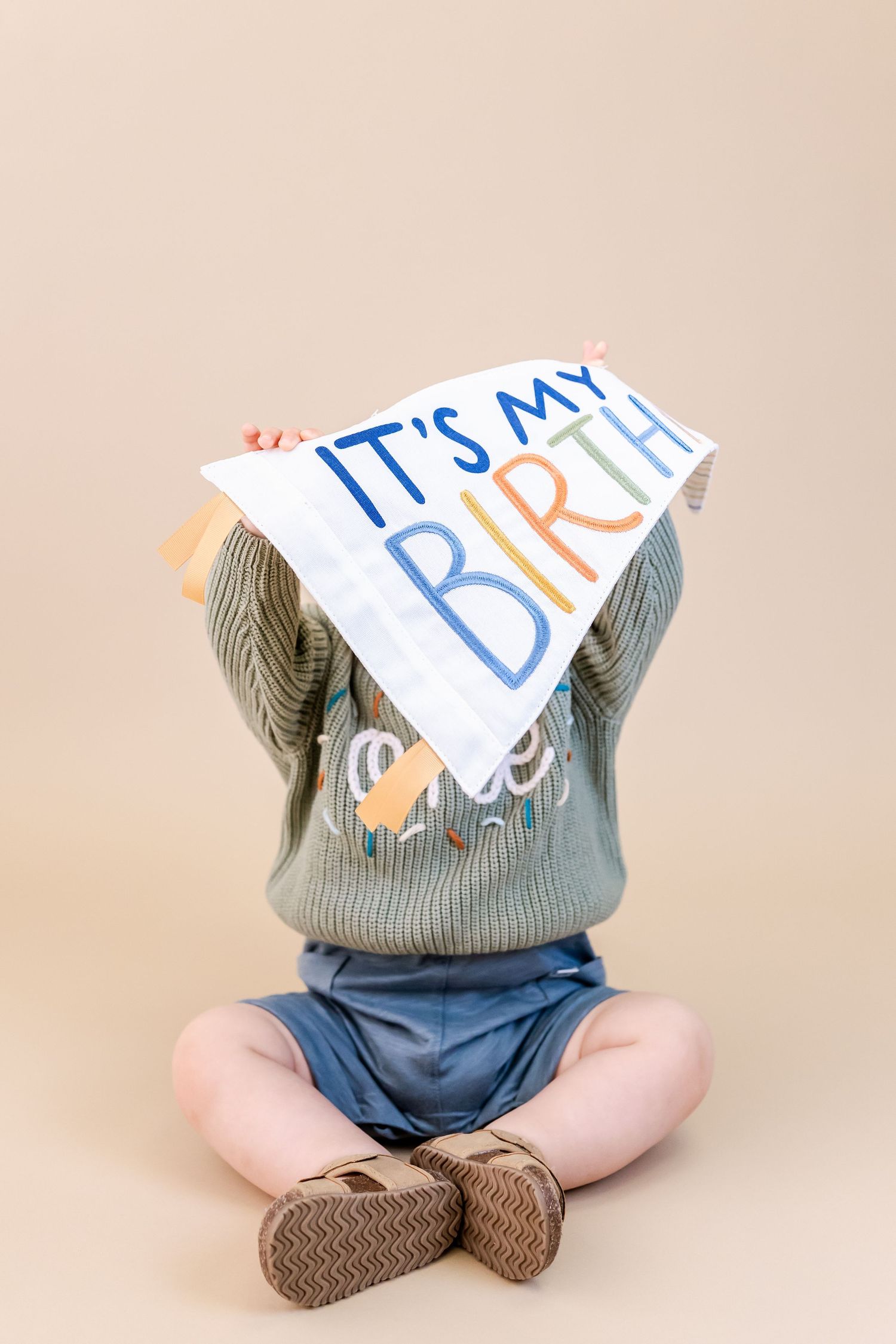 A baby in denim pants and gray shirt sits cross-legged on a beige background holding a sign.
