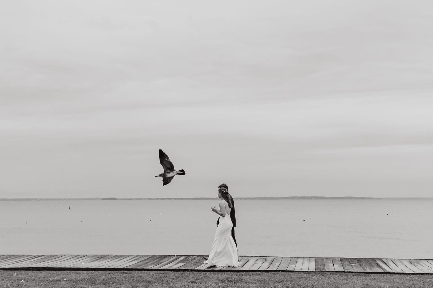 Black and white wedding photos capture a bride walking along a scenic waterfront boardwalk.