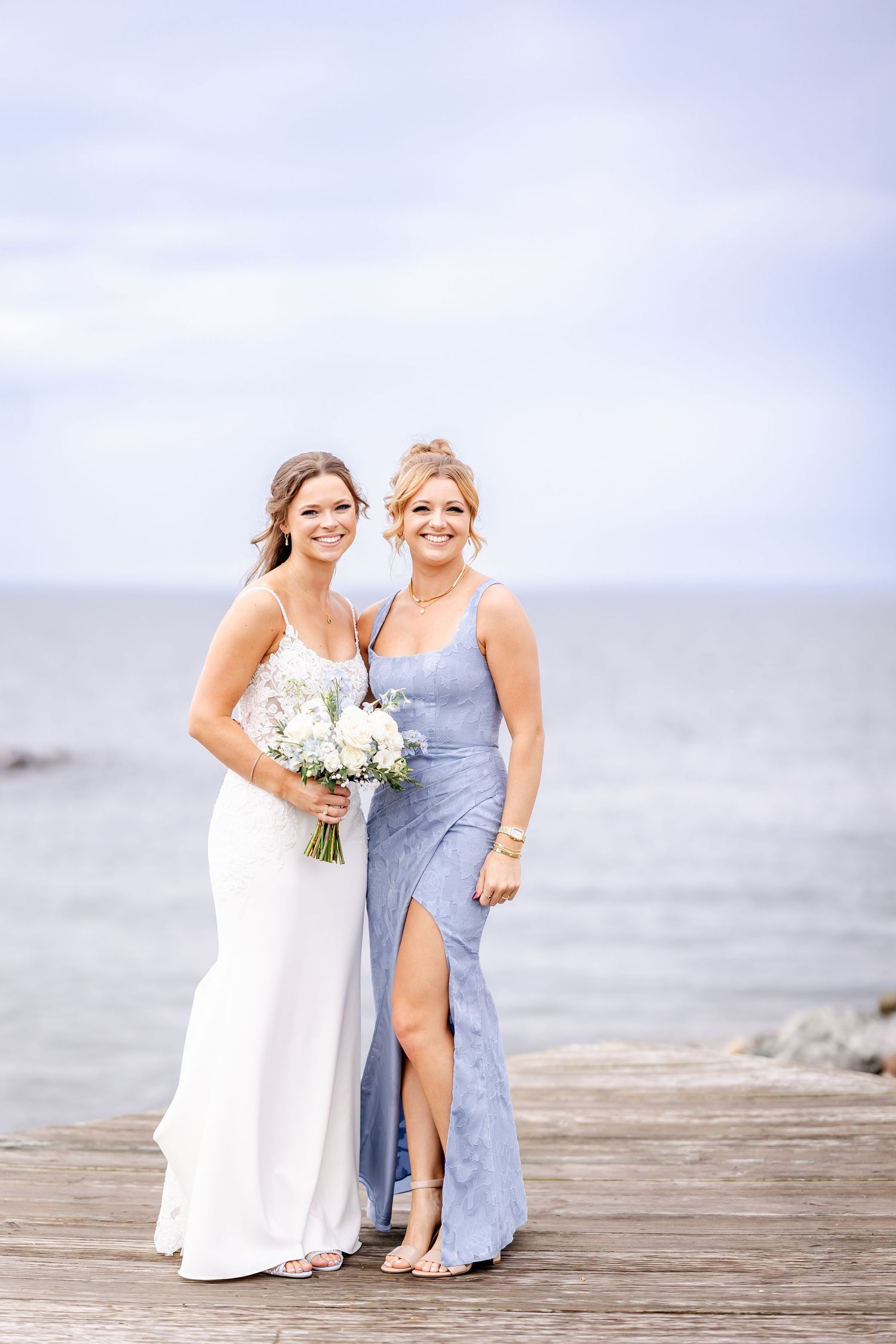 Two people in formal attire posing together on a wooden dock by the ocean.
