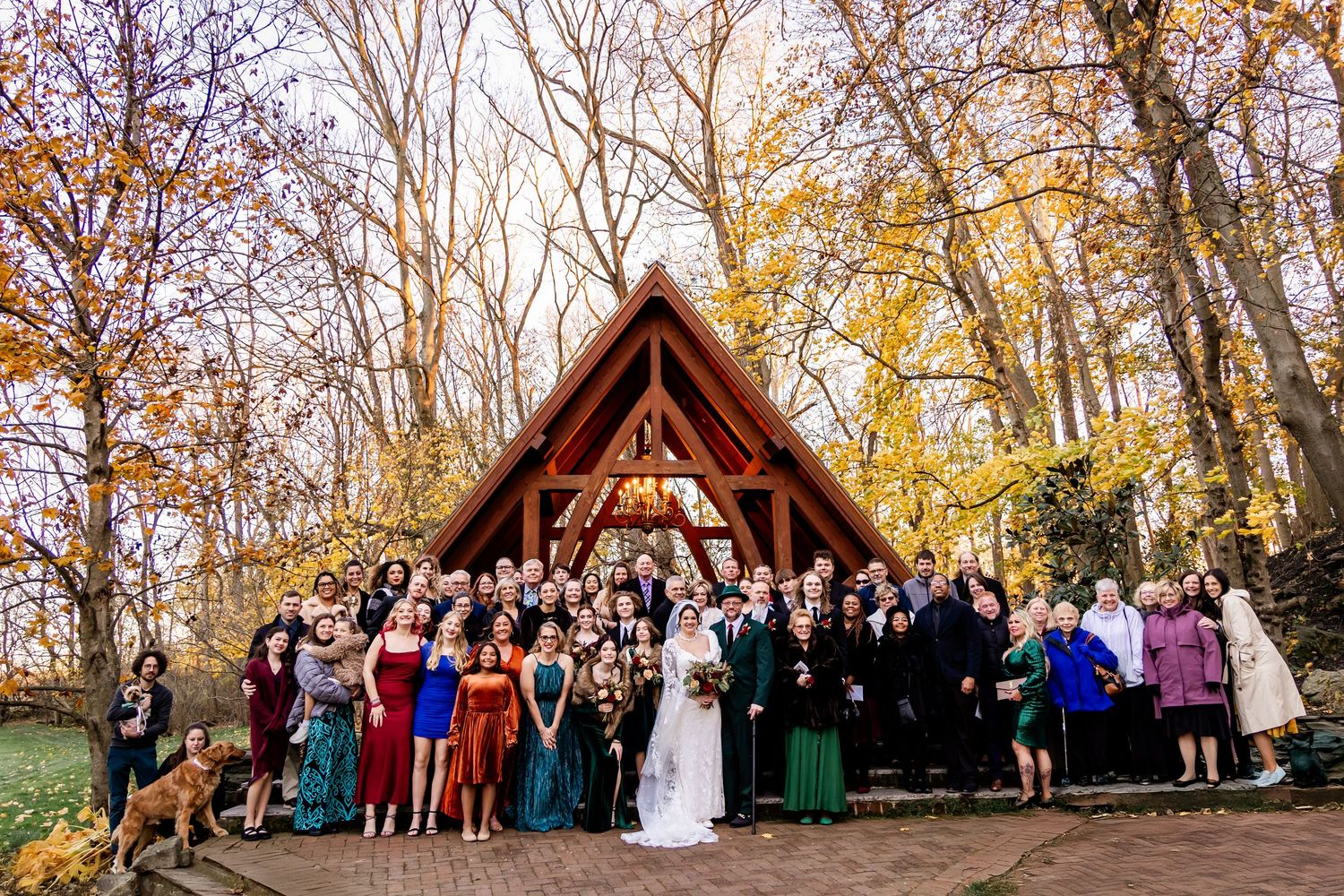 Large wedding party gathered outdoors under autumn trees near rustic wooden gazebo.