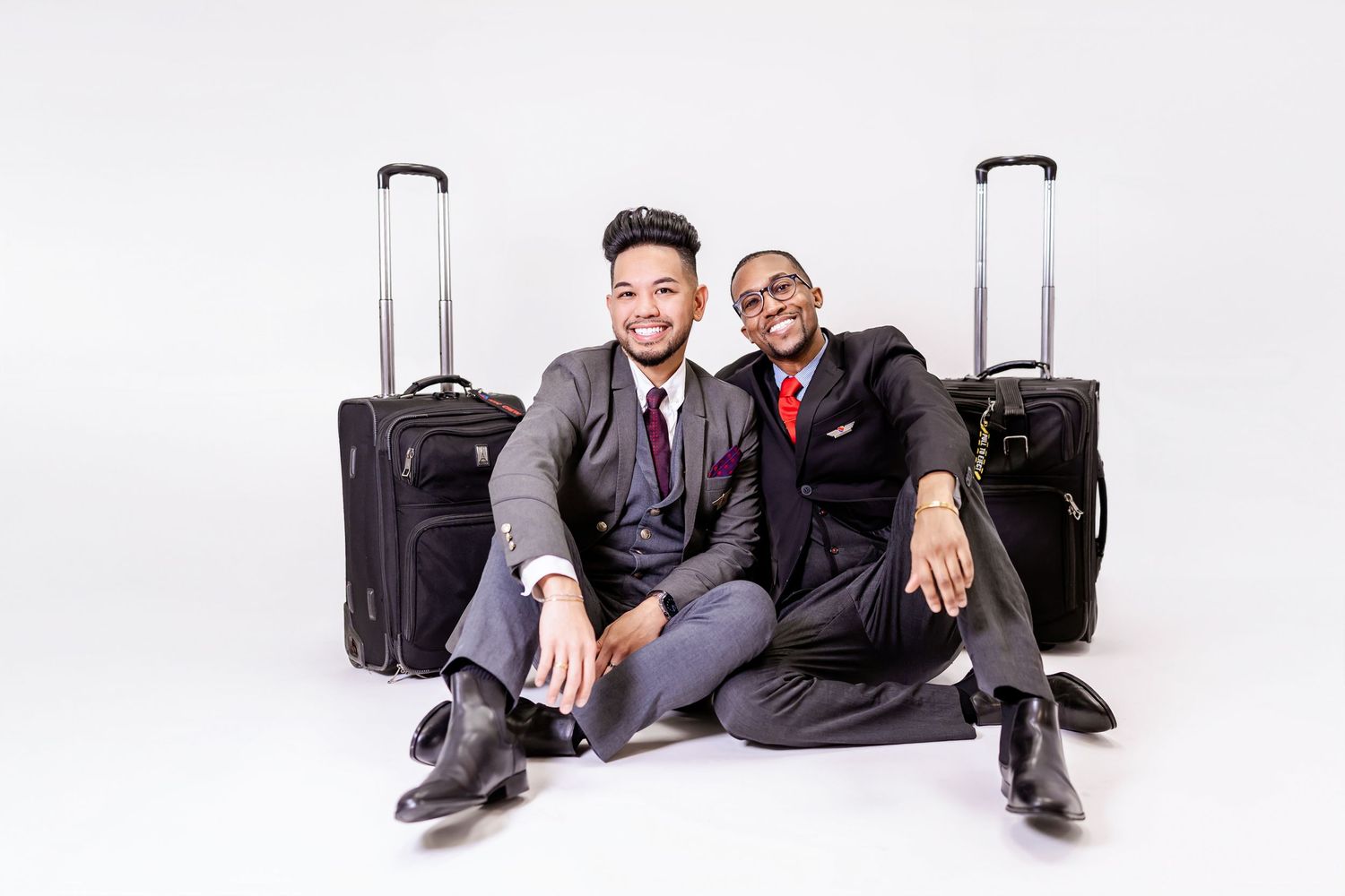 Two business travelers in suits sitting on floor with luggage trolleys against white background.