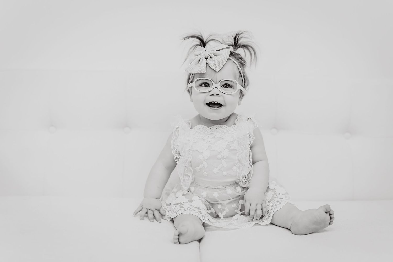 A baby in light dress and bow smiles against a simple grey backdrop.