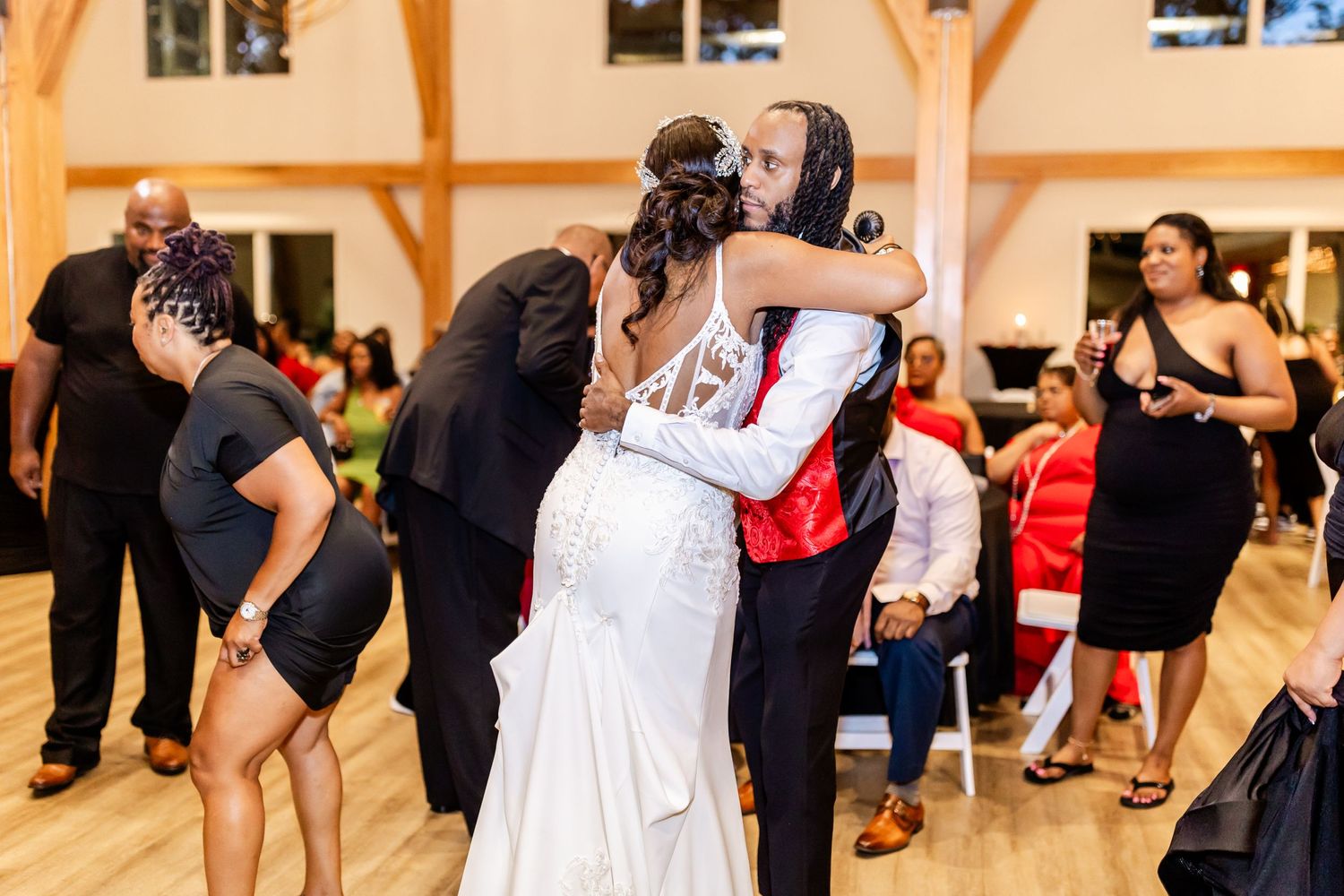 Wedding reception guests sharing embraces on a dance floor with wooden beams overhead.