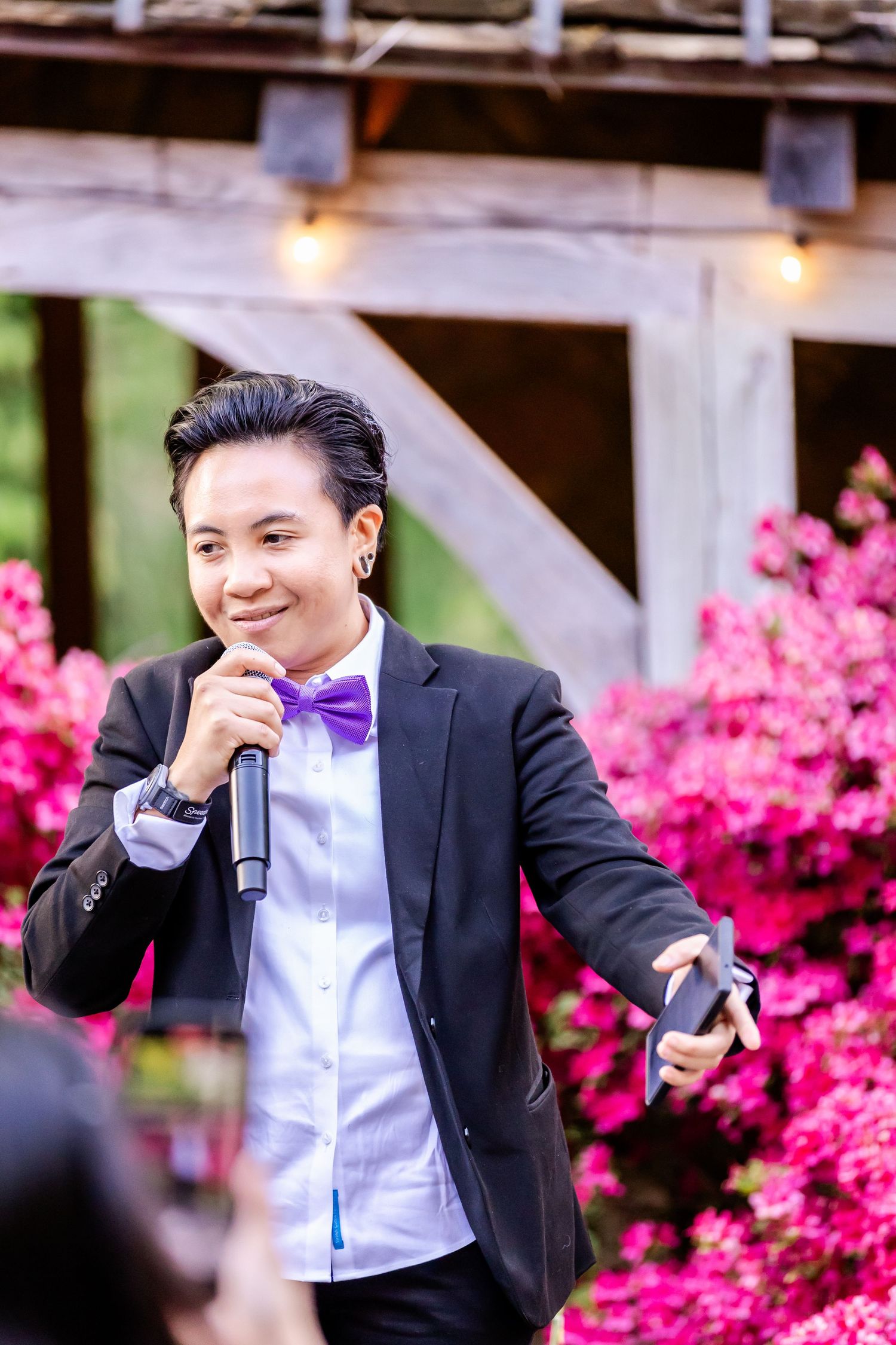 A performer in a purple bowtie and black jacket sings into a microphone outdoors.
