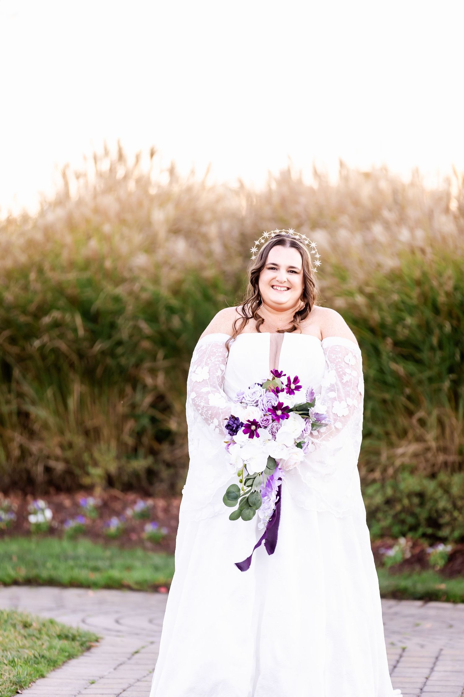 Bride in white lace dress holds purple and white bouquet against natural backdrop.