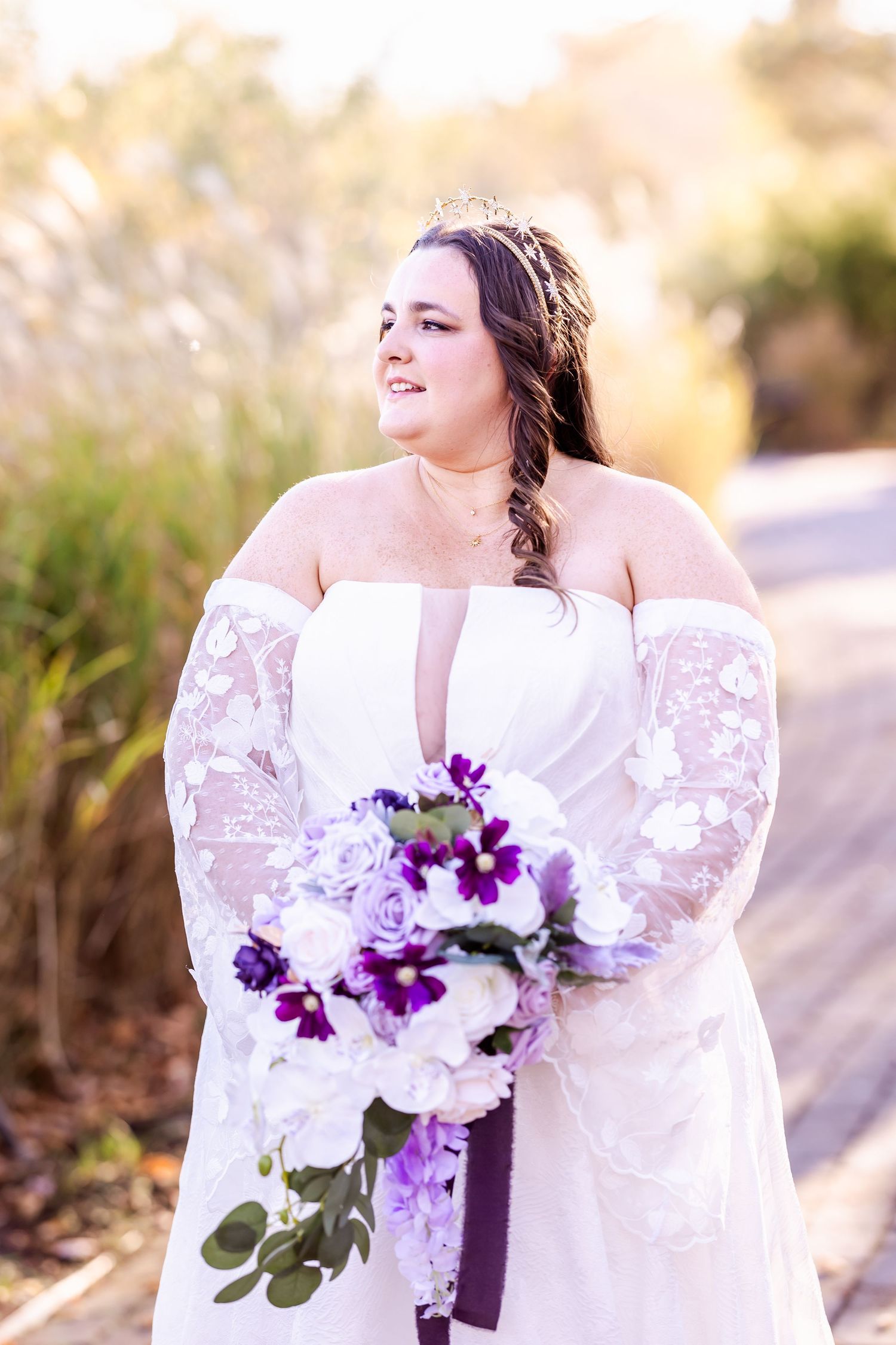 Close-up of bride holding purple and white floral bouquet in lace-sleeved wedding gown.