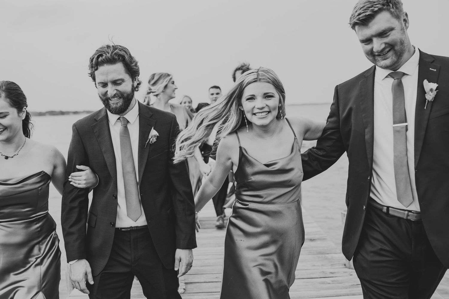 A happy wedding party walks together on a wooden dock wearing formal attire in black and white.