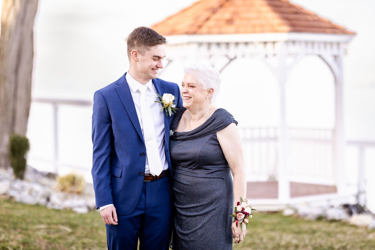 A couple in formal wedding attire embraces on a waterfront pier with a scenic backdrop.