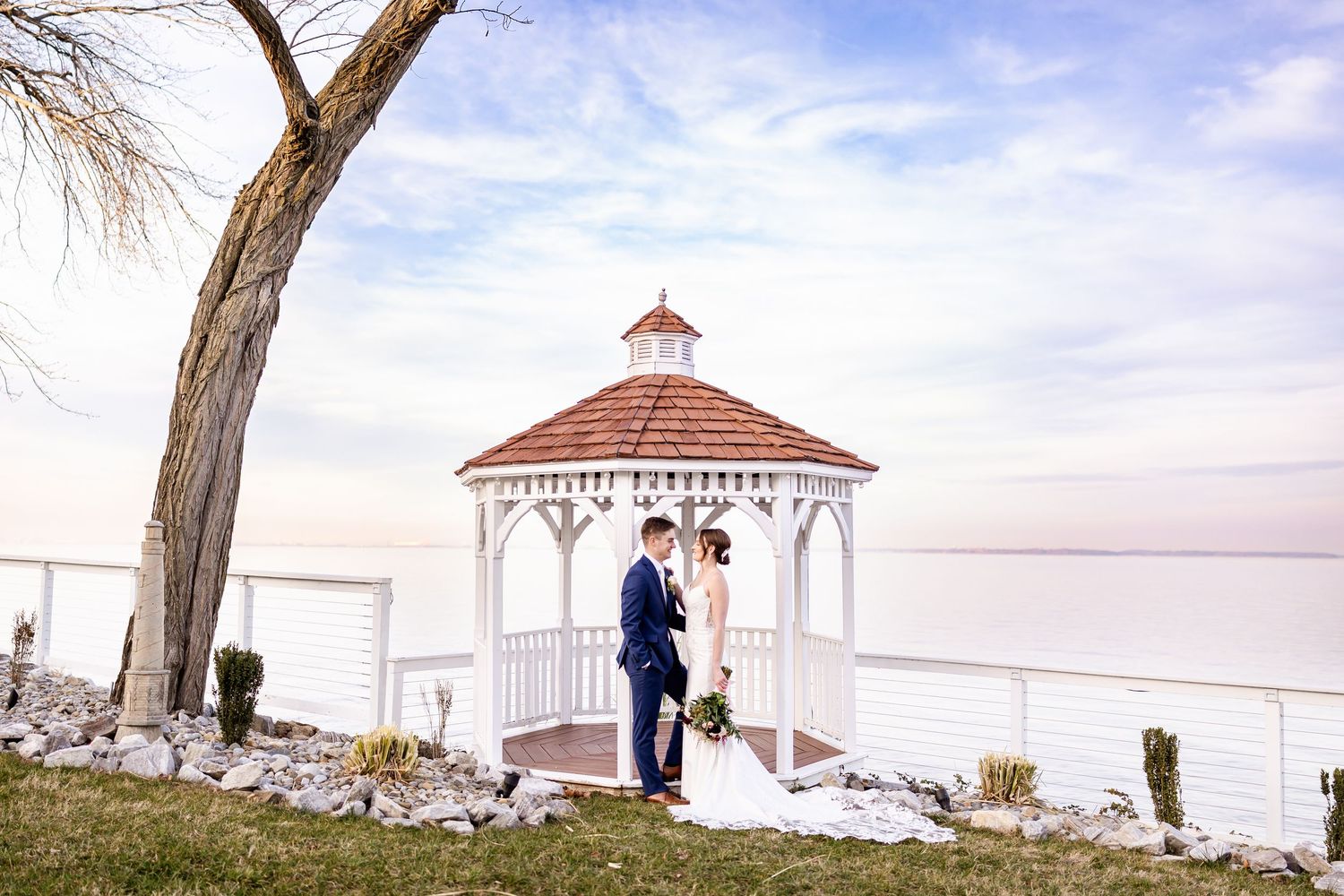 Scenic wedding photos taken near a white gazebo overlooking the water at sunset.