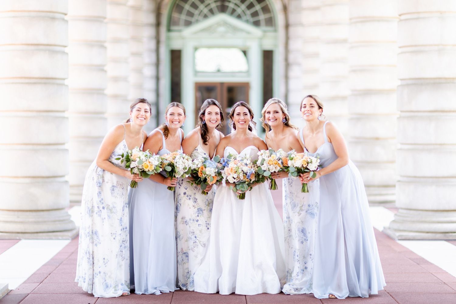 A bridal party in light blue and white dresses poses together holding pastel floral bouquets outside a classic building entrance.