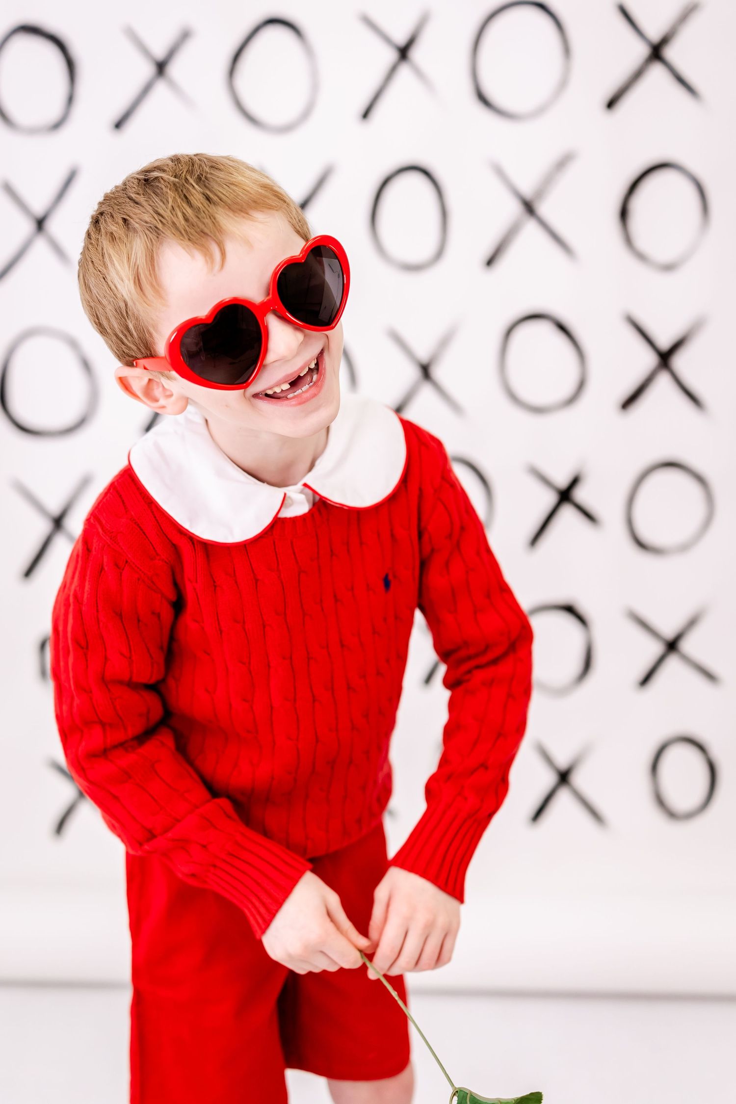 A happy child wearing a red sweater and heart-shaped sunglasses poses against a backdrop with XO pattern.