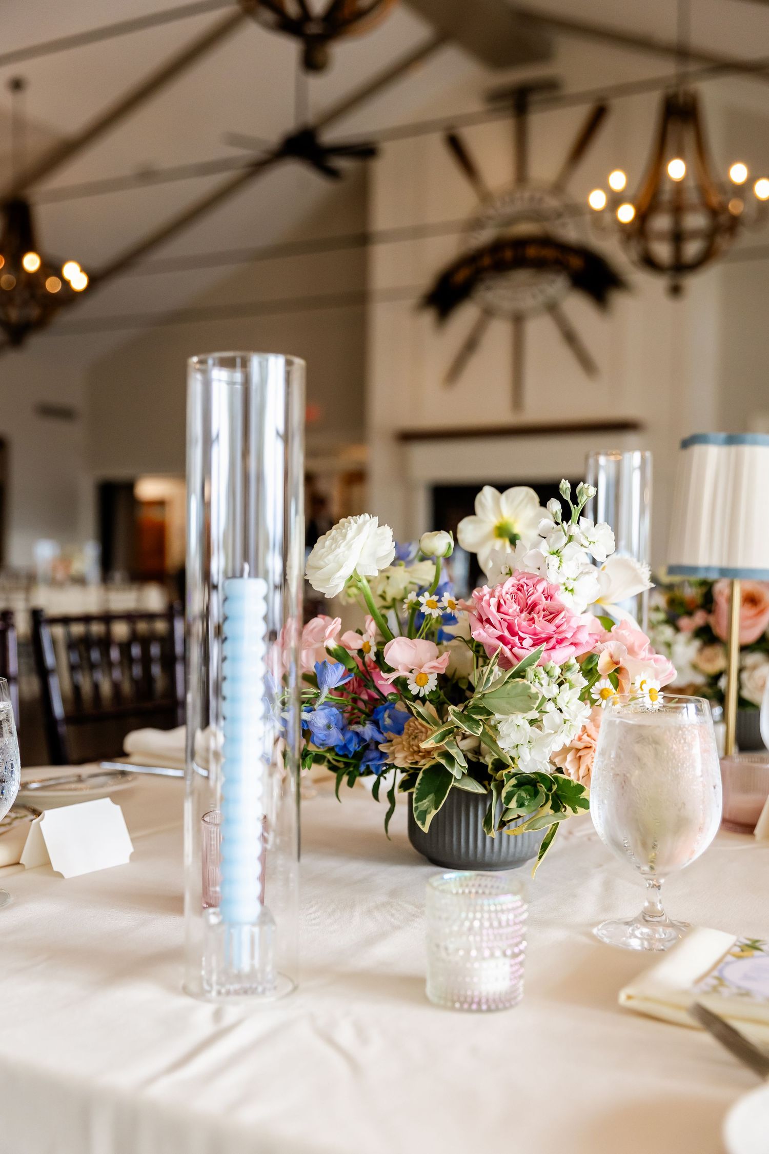 Wedding reception venue with white tablecloths, floral arrangements, and decorative lighting fixtures.