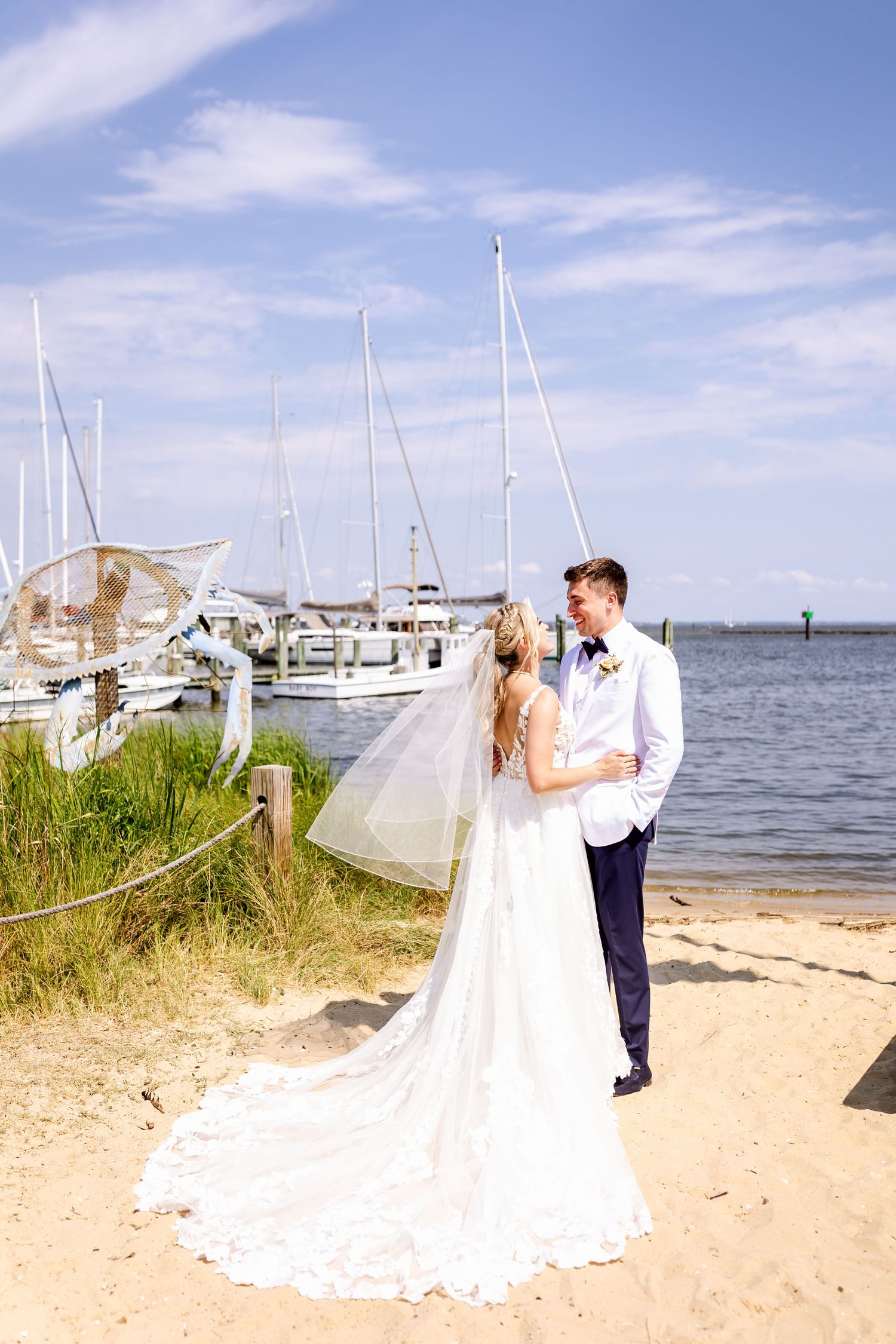 A wedding couple embraces on a sandy beach beside a marina with boats and blue sky in the background.