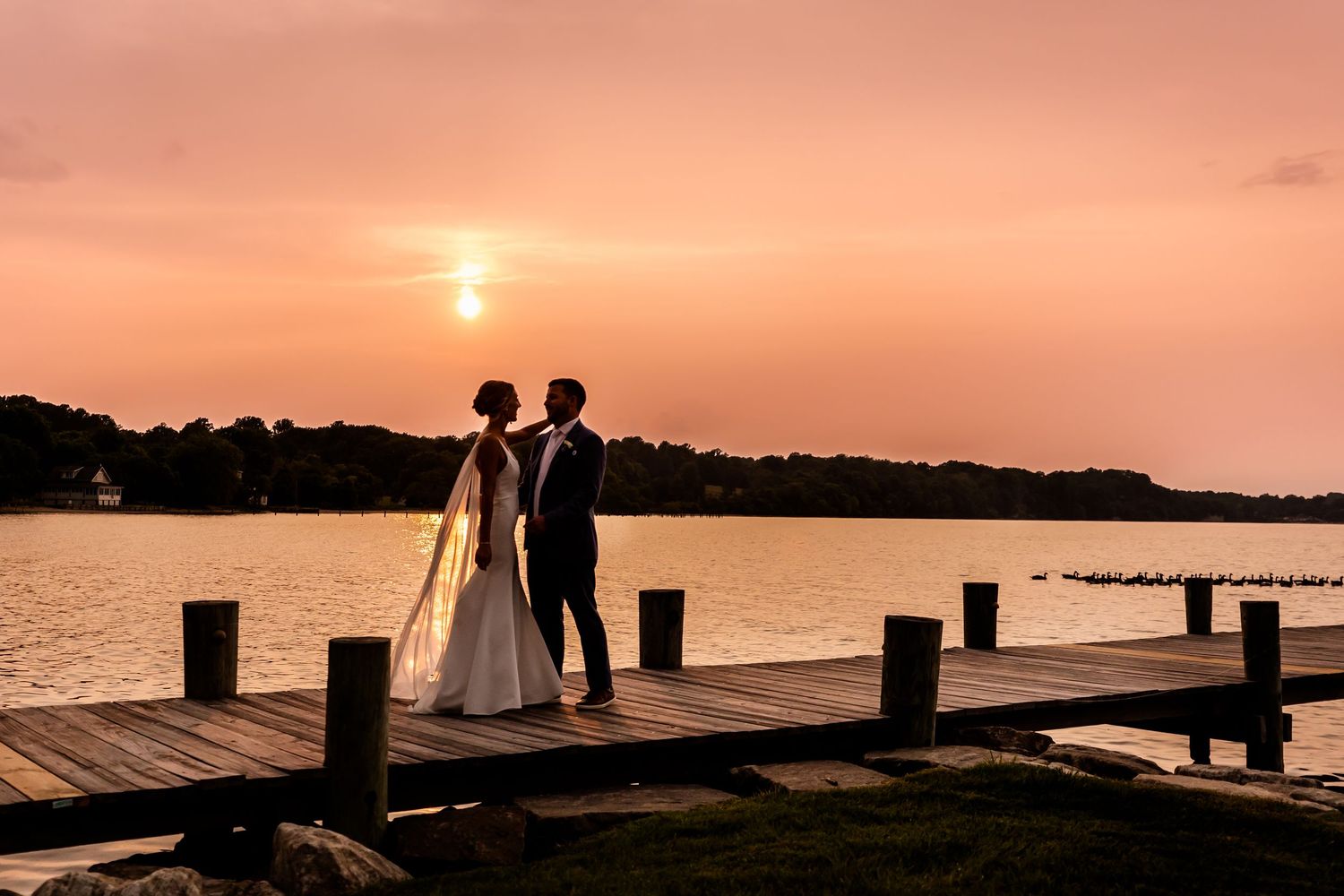 Silhouetted wedding couple stands on wooden pier during orange sunset over tranquil lake.
