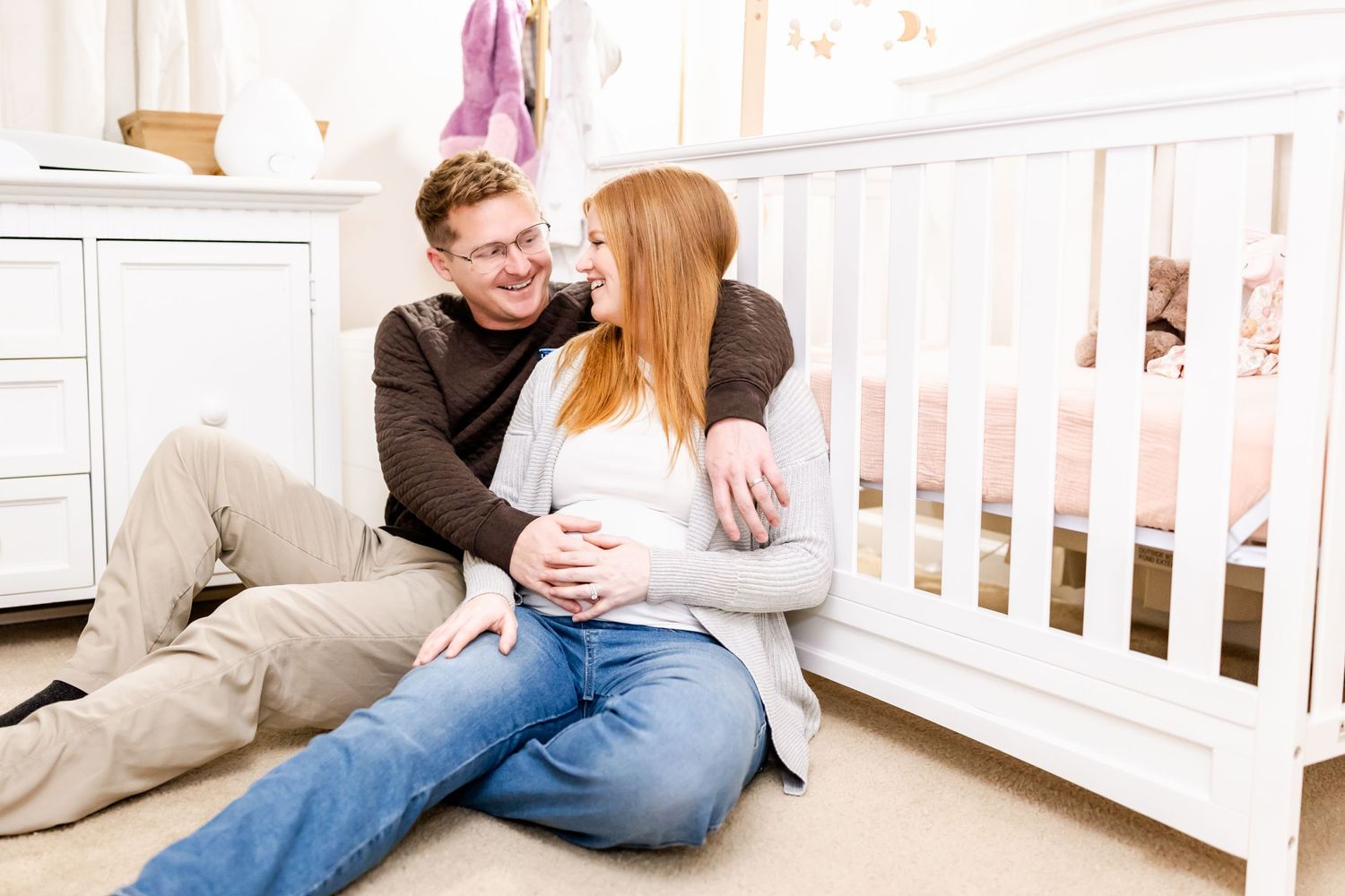 Expecting couple sits together on floor next to white crib in nursery sharing intimate moment.