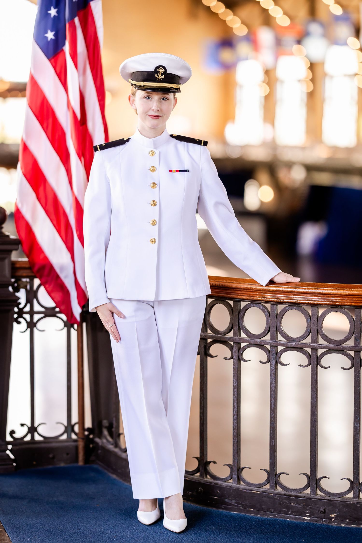 A naval officer in dress whites stands next to an American flag and ornate railing inside an elegant venue.