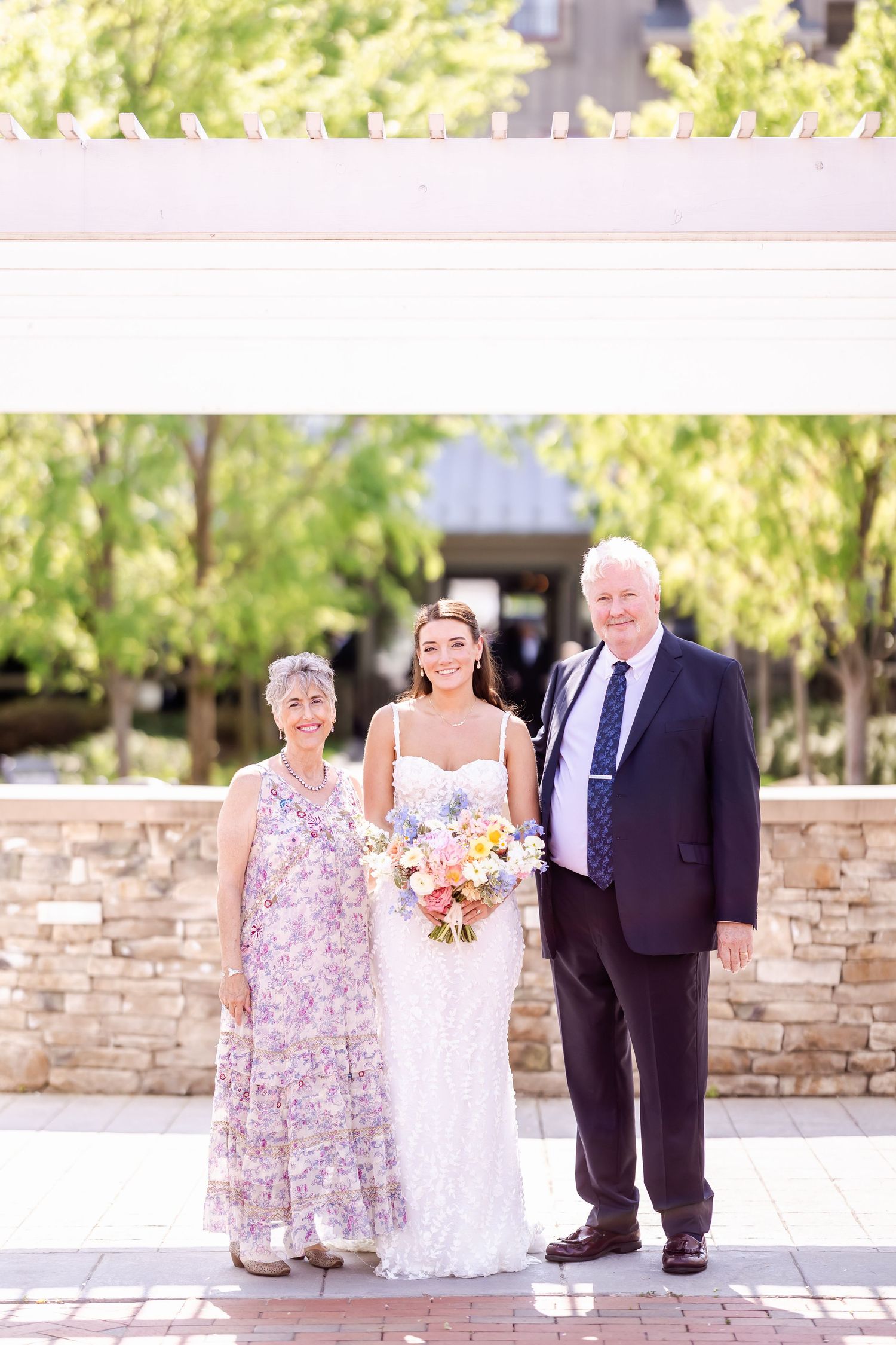 Wedding party members pose outdoors against a stone wall backdrop with floral bouquets.