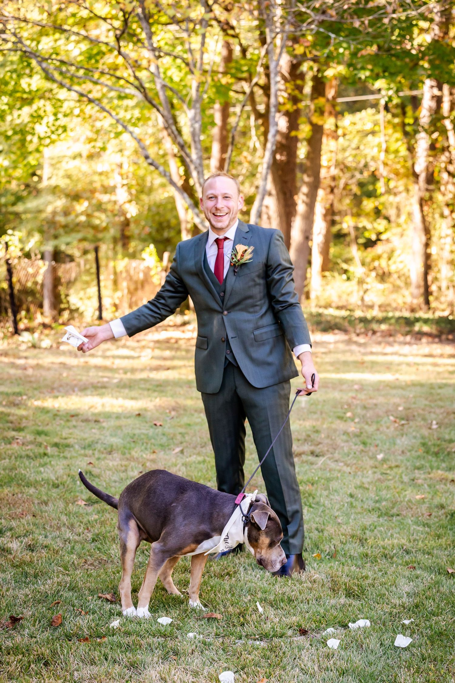 A groom in a dark suit plays with a dog on a grassy lawn during an outdoor wedding photoshoot.