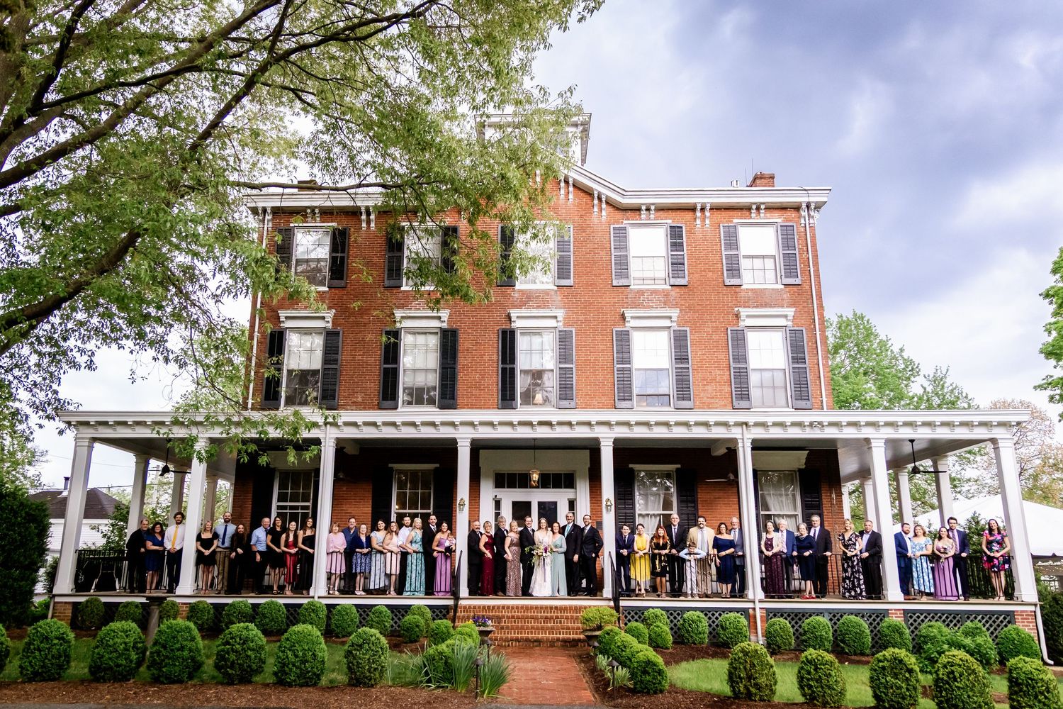 Historic red brick mansion with white columns and wraparound porch surrounded by lush green landscaping.