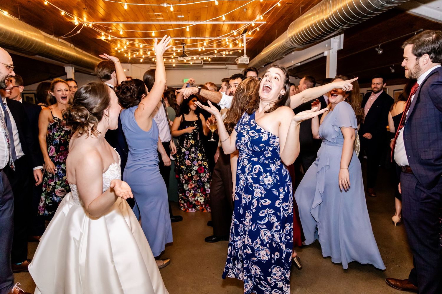 Wedding guests dance and celebrate on a rustic barn dance floor illuminated by warm string lights overhead.