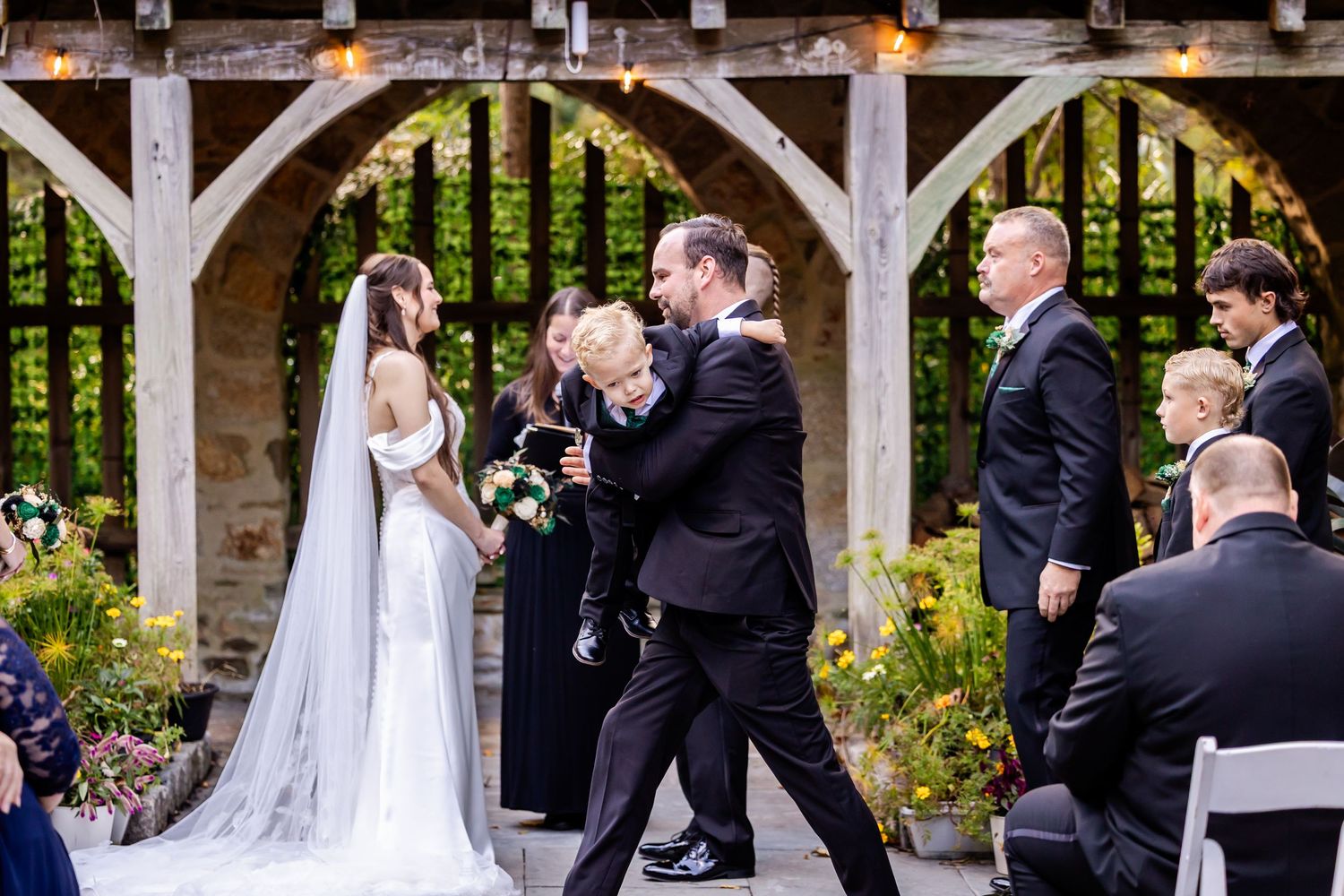 A wedding party poses under an elegant outdoor stone pergola during a formal ceremony.