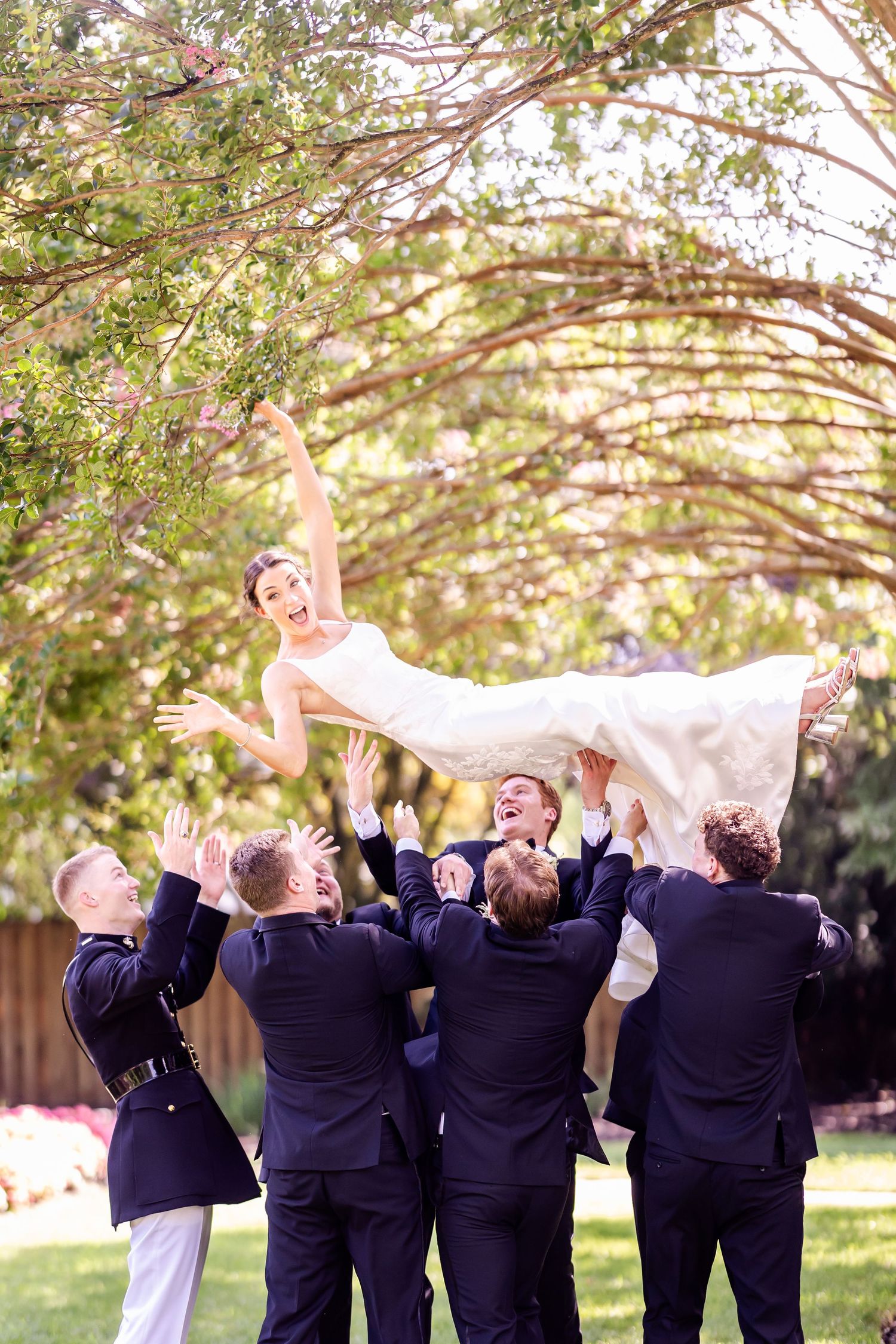 Wedding party lifts bride in white dress during playful outdoor photo session.