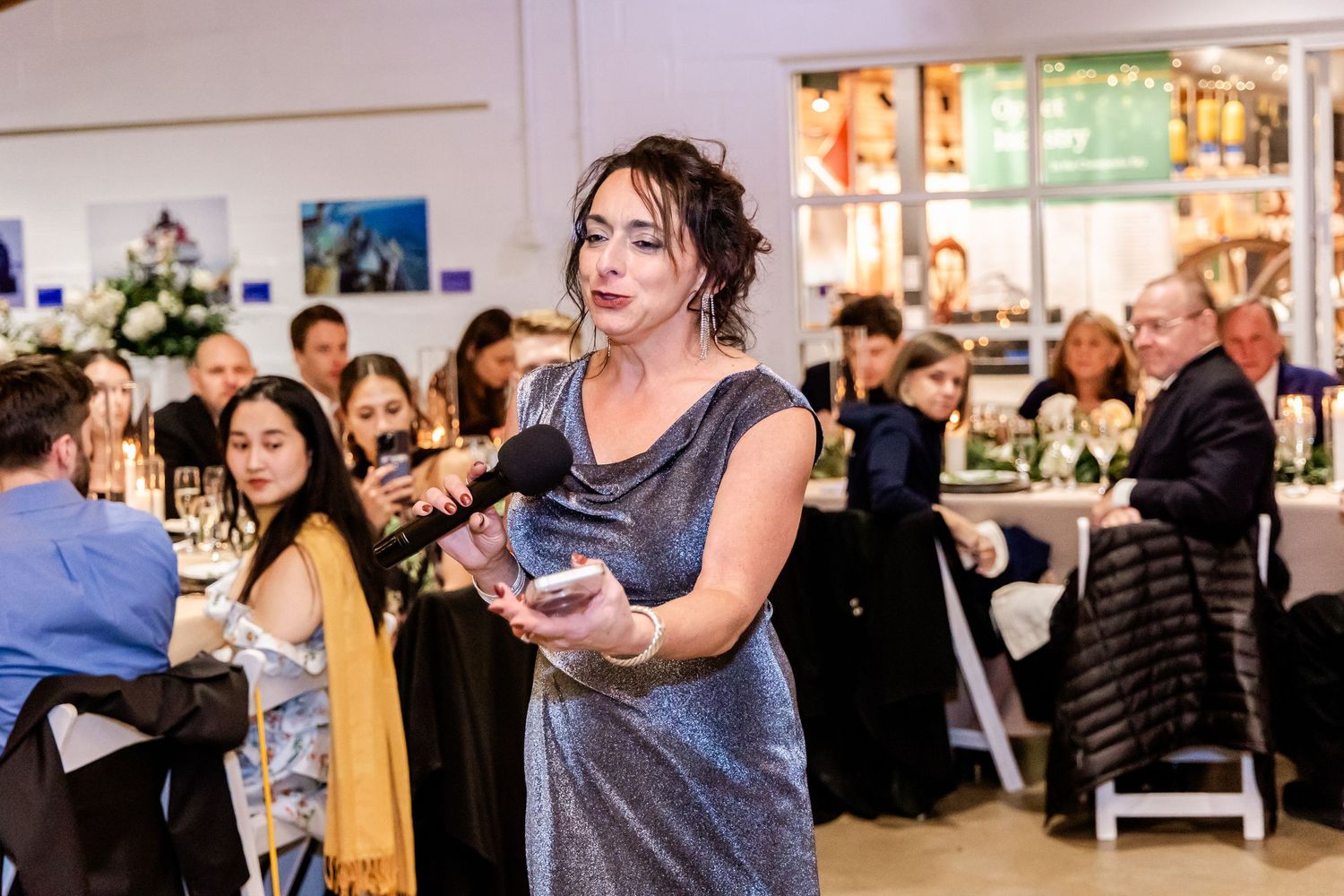 Woman in silver dress holds award while speaking at an elegant formal gala dinner event.